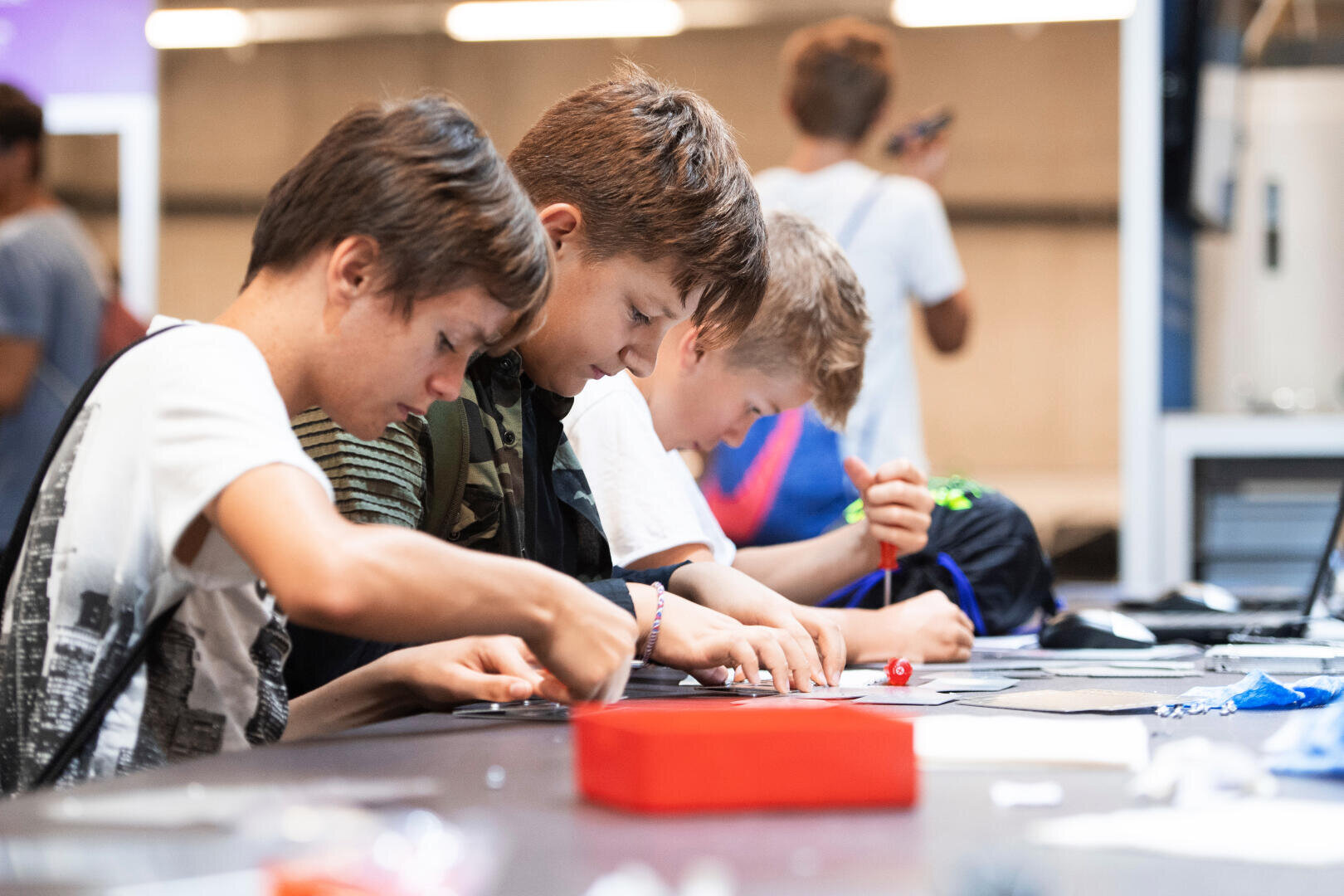 Drei Jungen sitzen nebeneinander an einem Tisch und arbeiten konzentriert an der Montage kleiner elektronischer oder handwerklicher Komponenten, während im Hintergrund weitere Kinder und Werkstattmaterialien zu sehen sind.