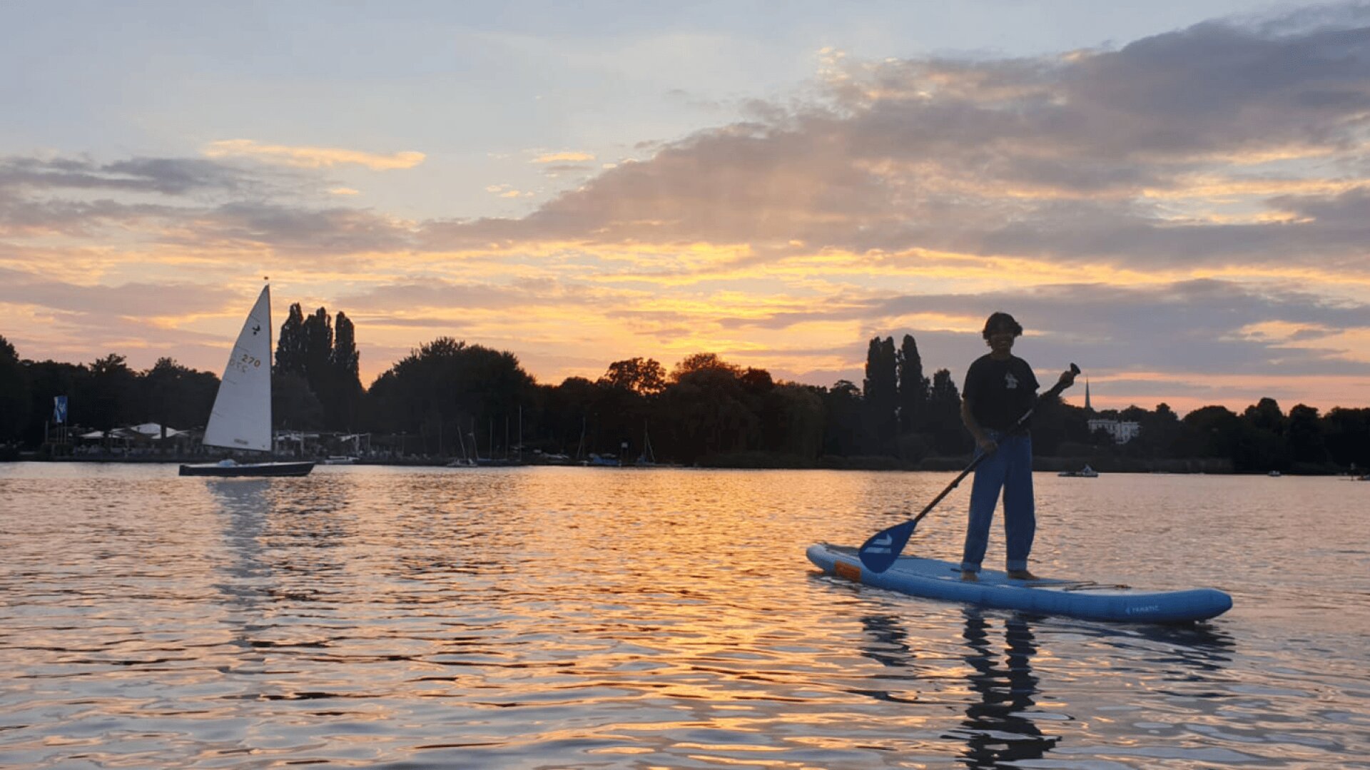 Eine Person paddelt auf einem ruhigen See bei Sonnenuntergang, mit einem Segelboot im Hintergrund und Bäumen, die sich gegen den bunten Himmel abheben.