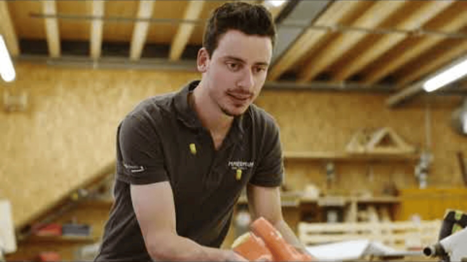 Ein Mann in einem grauen Poloshirt arbeitet in einer Holzwerkstatt und hantiert mit einem orangefarbenen Gegenstand auf einer Werkbank. Im Hintergrund sind Holzbalken und Tischlerwerkzeuge zu sehen.