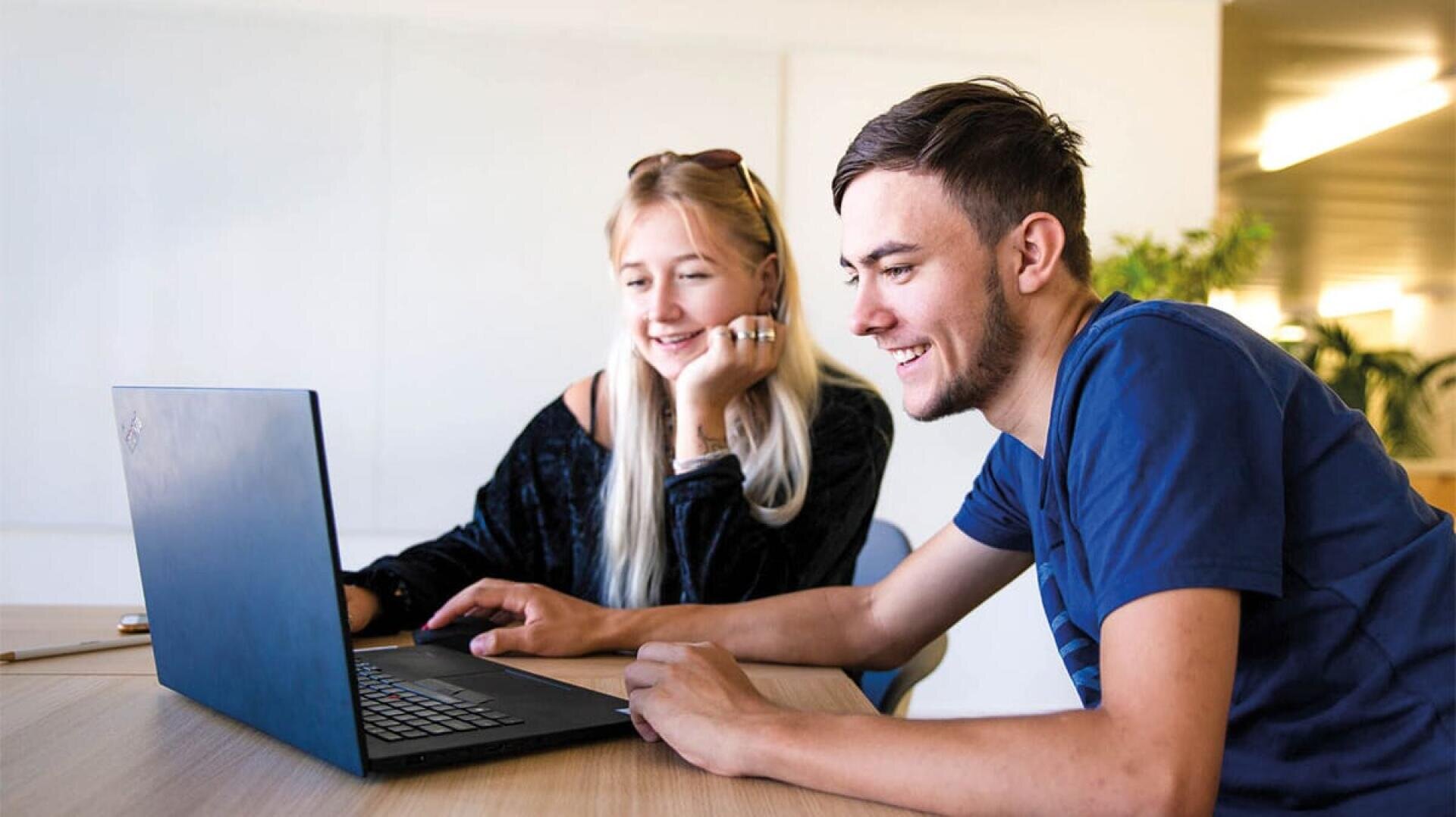 In einem hellen, modernen Raum sitzen zwei junge Erwachsene an einem Schreibtisch, lächeln und schauen gemeinsam auf einen Laptop-Bildschirm. Eine Person zeigt auf den Bildschirm, während die andere ihr Kinn auf ihre Hand stützt.