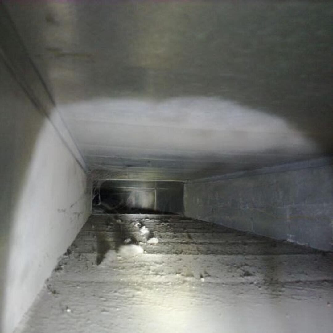 Ein staubiger Luftschacht im Inneren mit angesammeltem Schmutz und Schutt auf dem Boden, beleuchtet von einer Taschenlampe. Die Wände und die Decke erscheinen grau und metallisch.