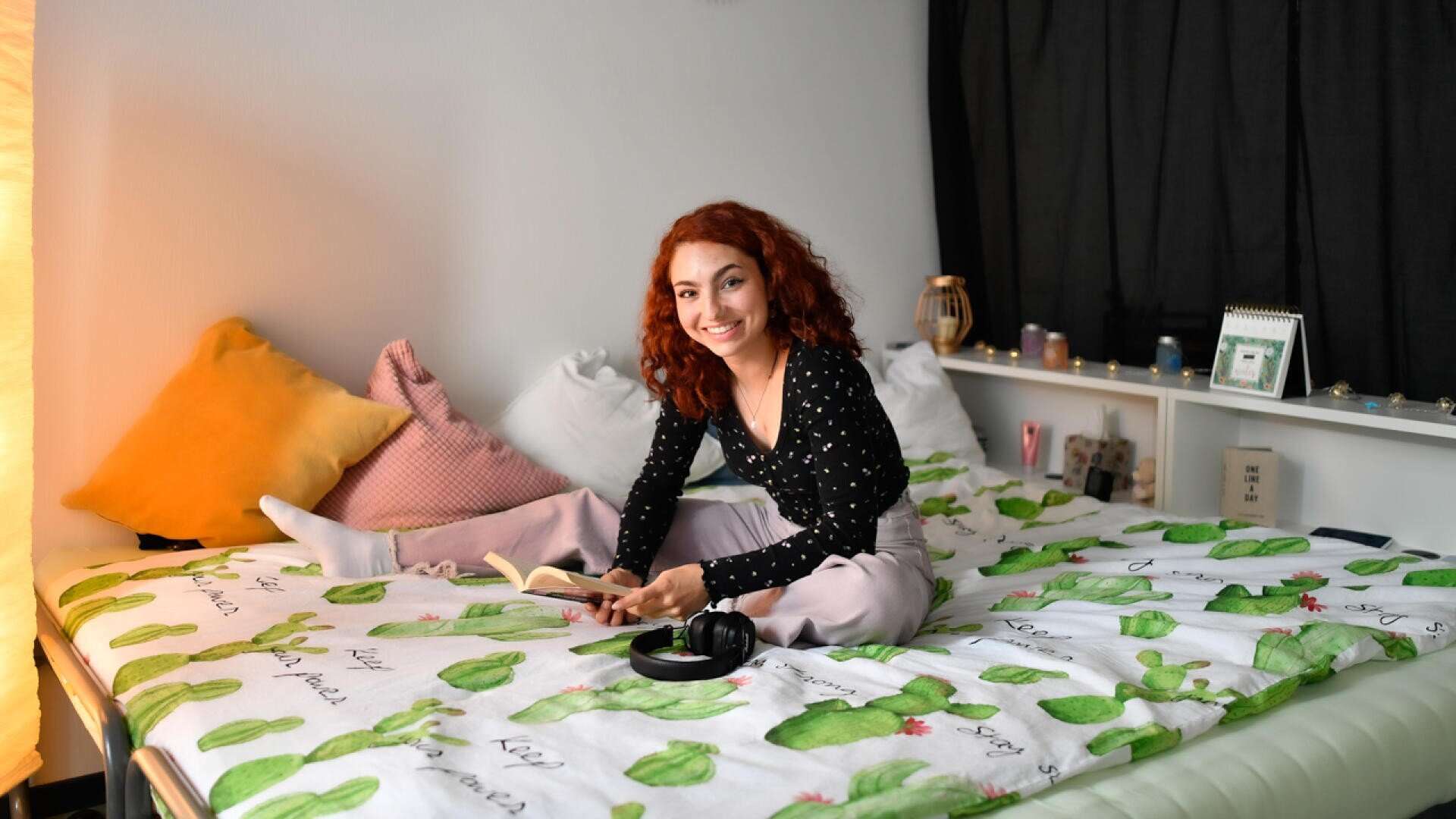Eine junge Frau mit roten Locken sitzt im Schneidersitz auf einem Bett mit einer kaktusgemusterten Decke und lächelt in die Kamera, während sie ein aufgeschlagenes Buch hält. Kopfhörer und ein Kissen liegen in der Nähe. Der Raum ist sanft beleuchtet.
