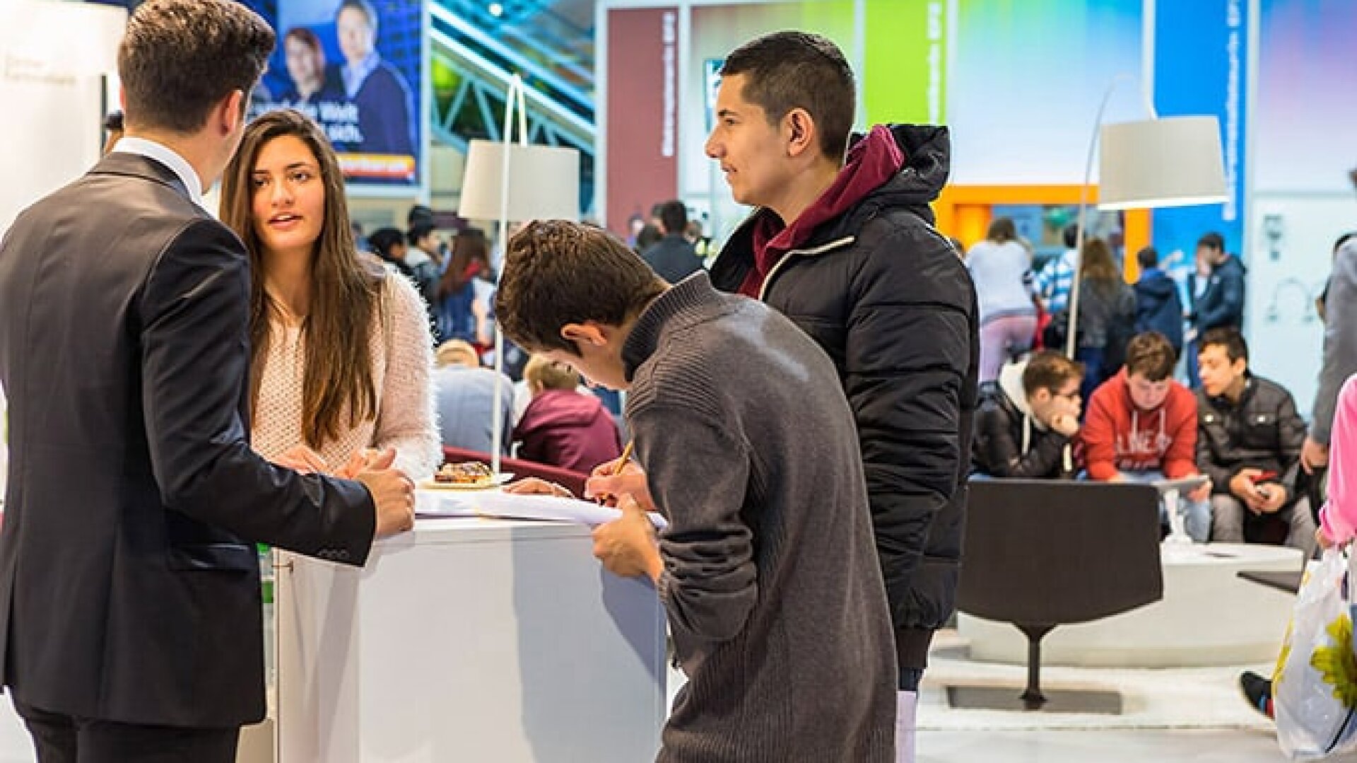 Eine Gruppe junger Erwachsener steht und unterhält sich auf einer belebten Veranstaltung oder Messe. Zwei Personen stehen sich gegenüber, während eine andere Person auf einem Klemmbrett schreibt. Der Hintergrund ist bunt, weitere Personen sitzen und gehen.