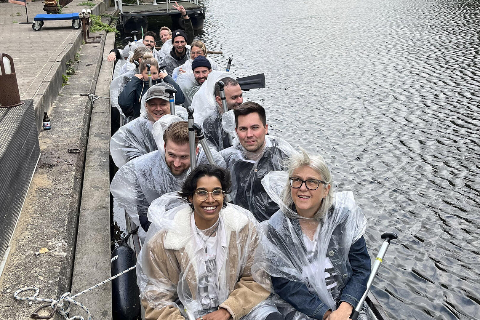 Eine Gruppe von Menschen in durchsichtigen Regenponchos sitzt eng zusammen in einem kleinen Boot auf einem Kanal und lächelt in die Kamera. Das Boot liegt an einer Betonkante angedockt. Neben ihnen ist das Wasser ruhig.