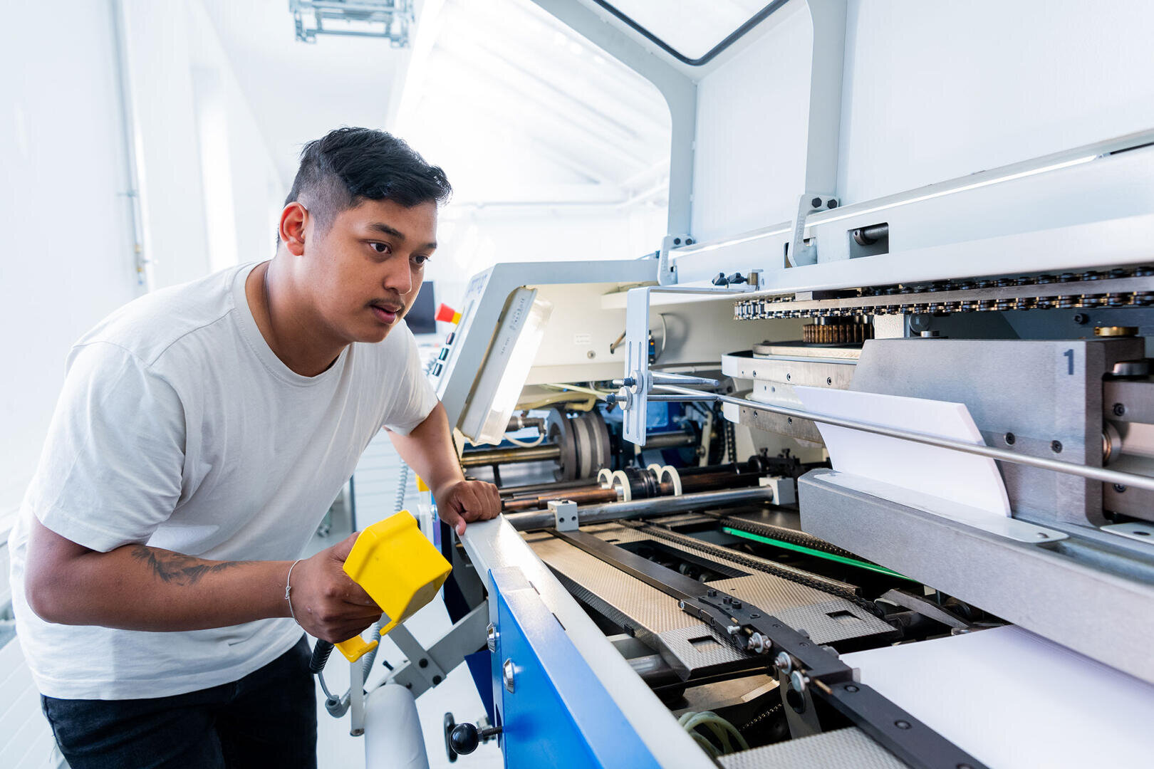 Ein Mann in einem weißen T-Shirt inspiziert in einer hell erleuchteten Industrieumgebung eine große, moderne Druckmaschine, in der sich Papierbögen befinden, und hält einen gelben Gegenstand in der Hand.