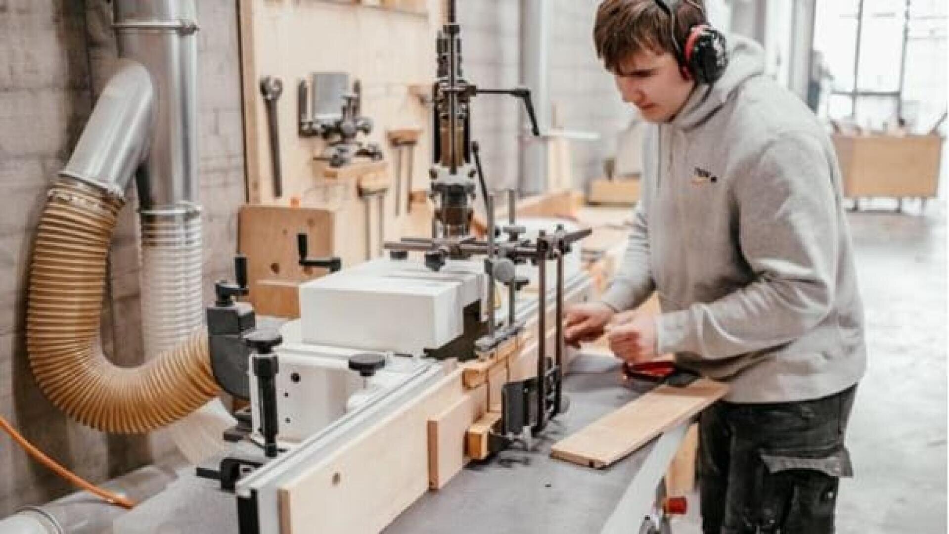 Eine Person, die einen Gehörschutz trägt, bedient eine Holzbearbeitungsmaschine in einer Werkstatt und führt ein Holzstück durch die Anlage. Auf der Werkbank und an den Wänden sind verschiedene Werkzeuge und Materialien zu sehen.