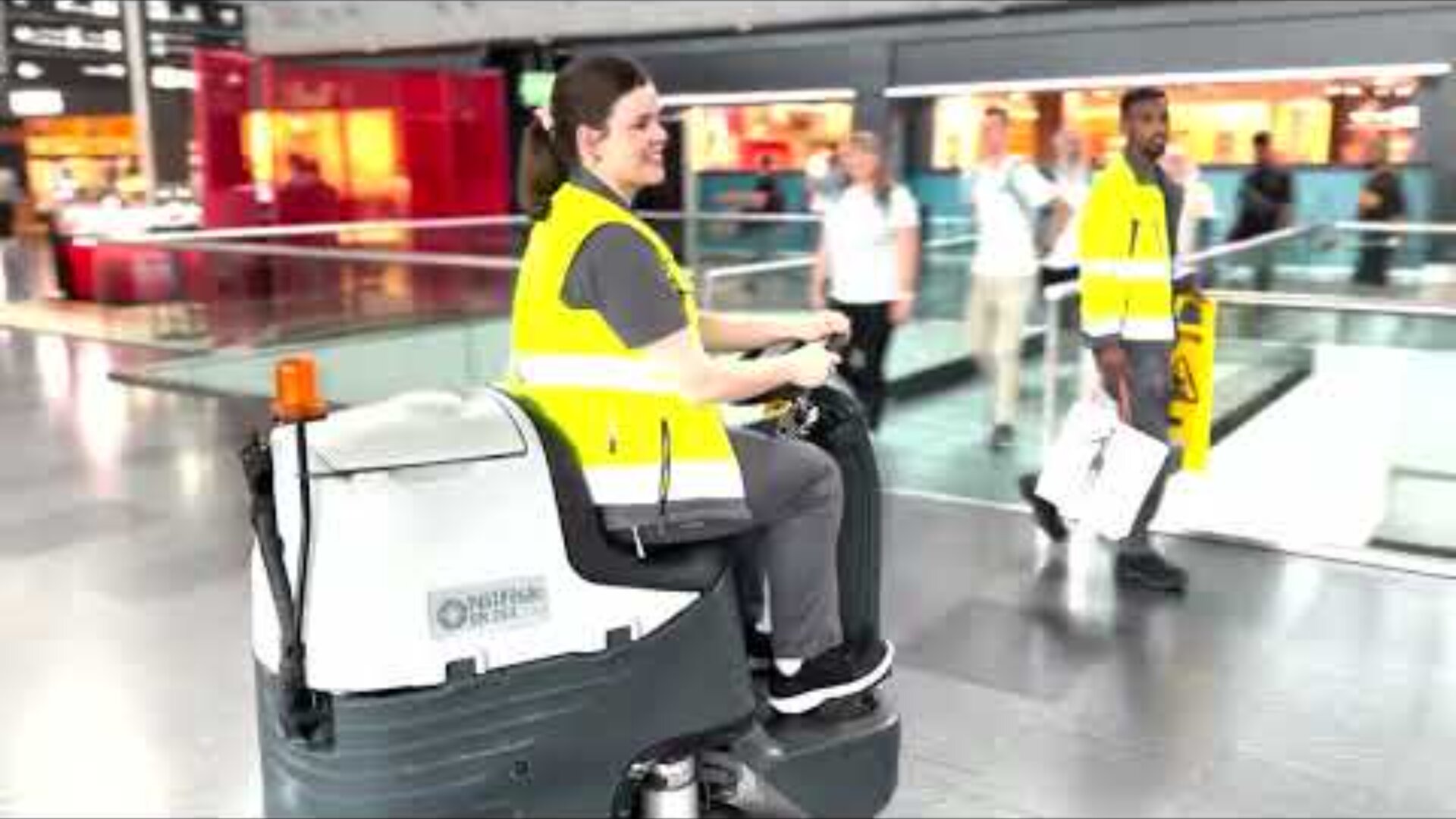 Ein lächelnder Arbeiter in einer gelben Sicherheitsweste bedient eine Bodenreinigungsmaschine in einem hellen, belebten Innenraum, während im Hintergrund andere Menschen vorbeigehen.