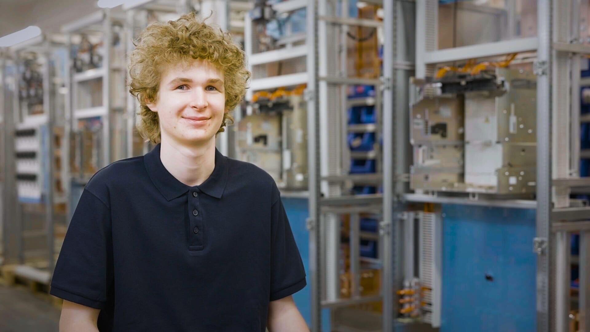 Eine junge Person mit lockigem Haar und schwarzem Polohemd steht lächelnd vor elektrischen Industriegeräten und Metallregalen in einer gut beleuchteten Arbeitsumgebung.