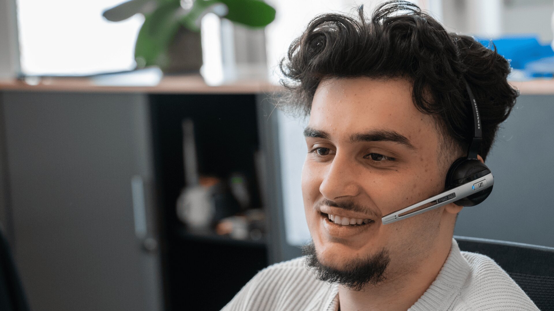 Ein junger Mann mit dunklem, lockigem Haar und Spitzbart lächelt, während er ein Headset-Mikrofon trägt und in einem Büro mit Regalen und einer Pflanze im Hintergrund sitzt.