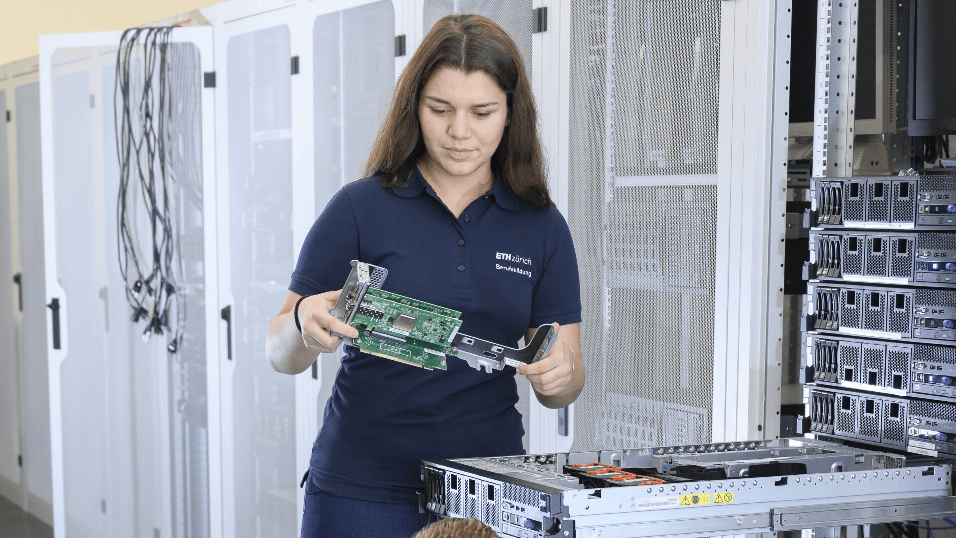 Eine Frau in einem marineblauen Polohemd prüft eine Computerplatine in einem Serverraum, der neben einem Regal mit offenen und gestapelten Servern steht.
