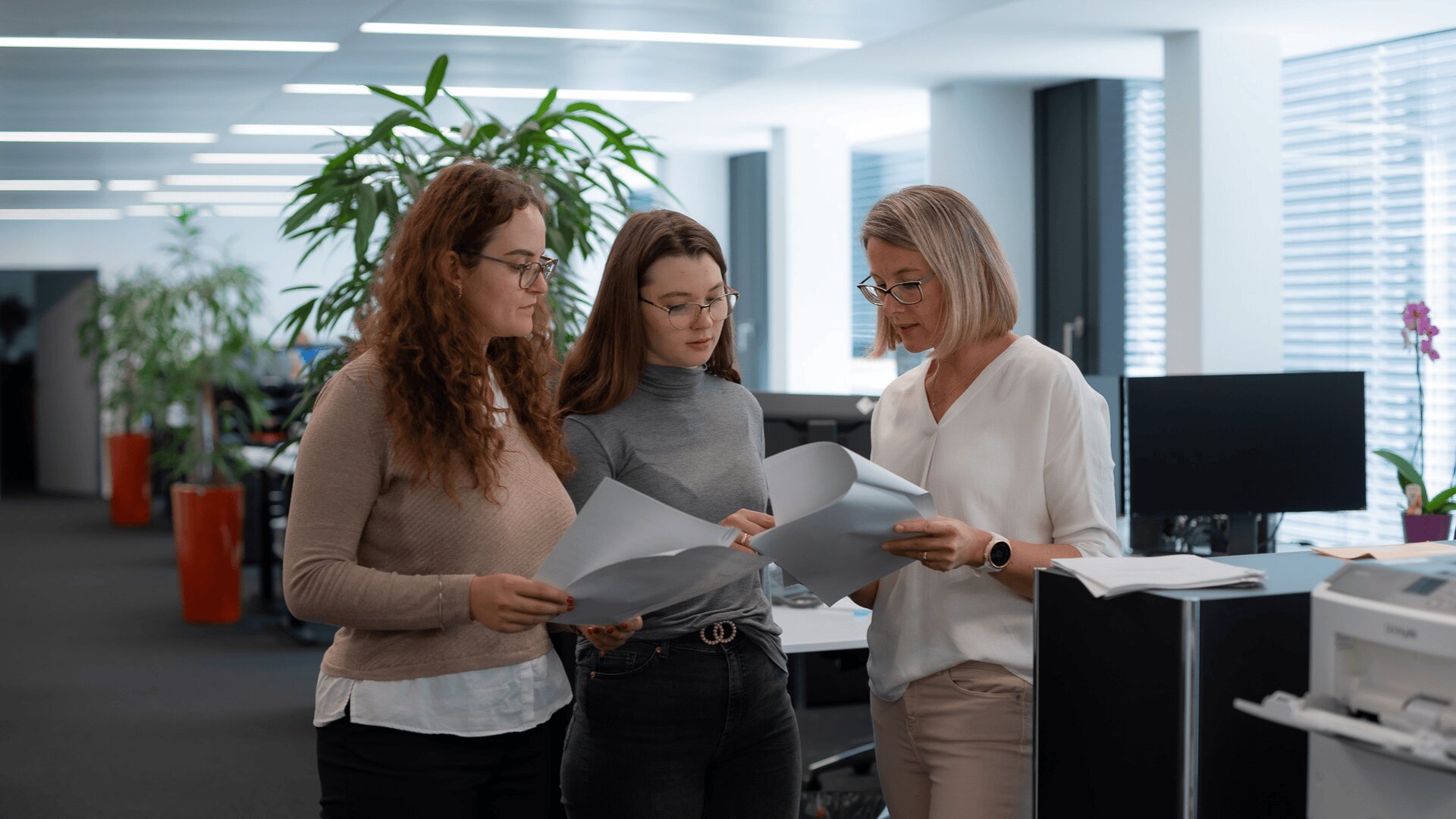 Drei Frauen stehen zusammen in einem modernen Büro und sehen sich Dokumente an. Zwei sind jung, haben lange Haare und Brillen, während die dritte, ältere Frau in Weiß, etwas erklärt. Computer, Pflanzen und natürliches Licht erfüllen den Arbeitsraum.