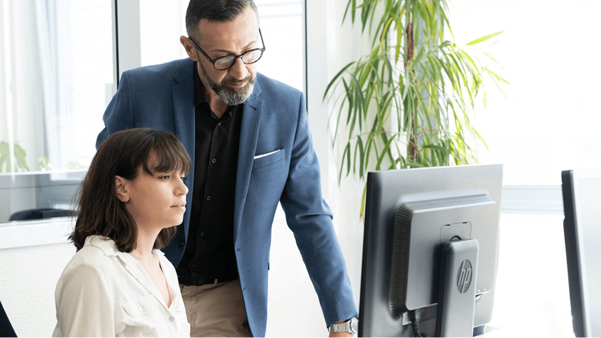 Ein Mann in einem blauen Blazer steht neben einer sitzenden Frau in einem weißen Hemd. Beide schauen auf einen Computerbildschirm in einem Büro mit großen Fenstern und einer Topfpflanze im Hintergrund.
