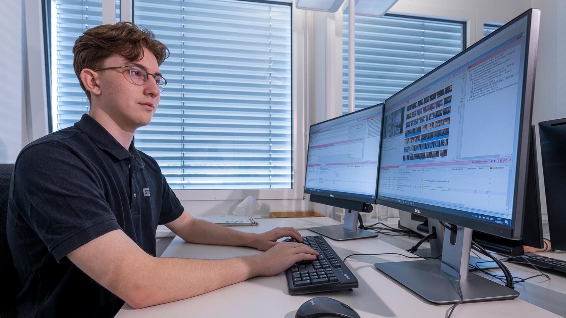 Eine Person mit Brille und schwarzem Polohemd sitzt an einem Schreibtisch mit Tastatur und Maus und arbeitet an zwei großen Computermonitoren, die Software und Dateien in einem hellen Büro mit Jalousien anzeigen.