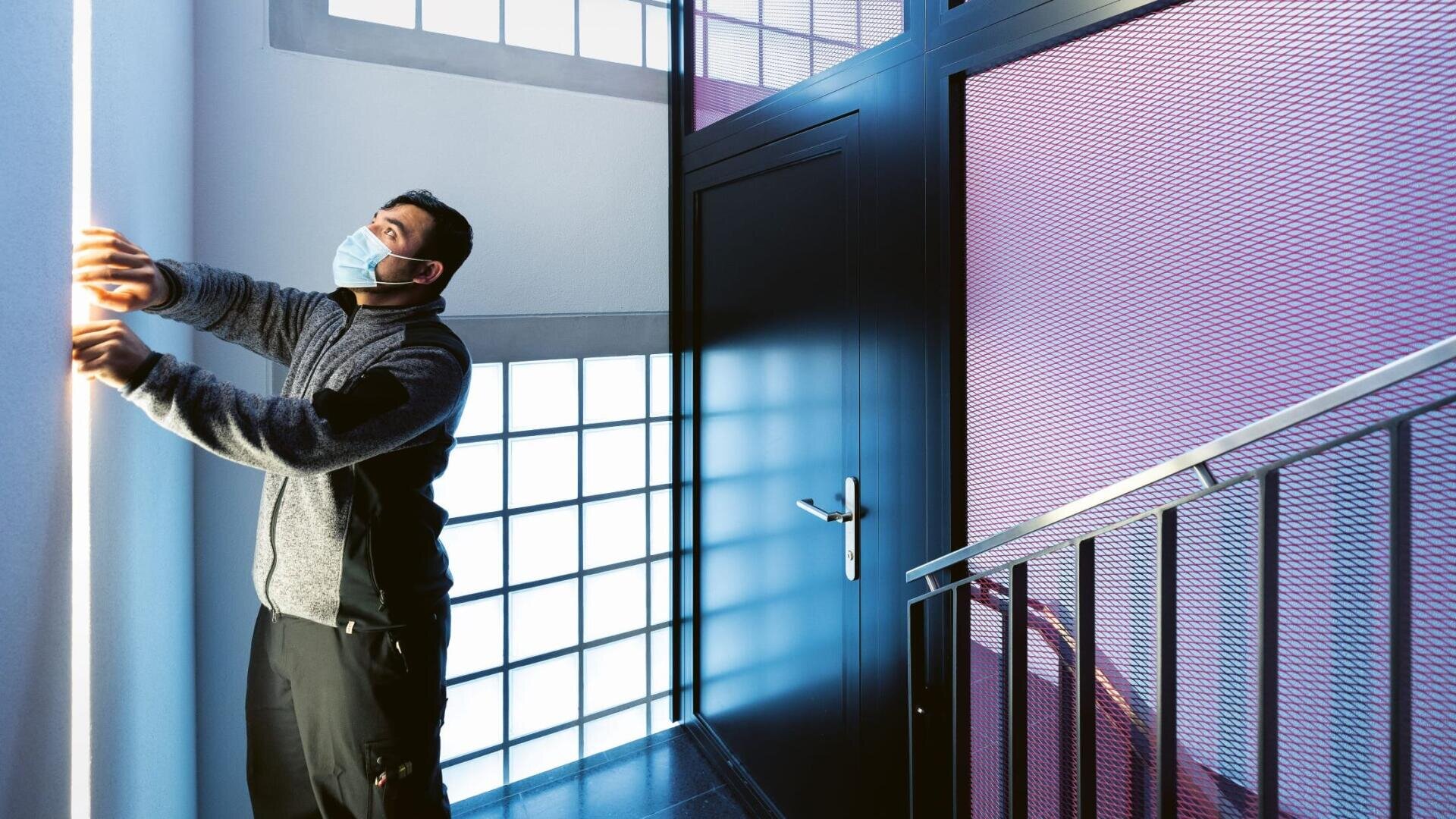 Eine Person mit einer Gesichtsmaske justiert ein vertikales Lichtband an einer Wand in einem modernen Treppenhaus, mit Glasbausteinen und einer Metalltür im Hintergrund.
