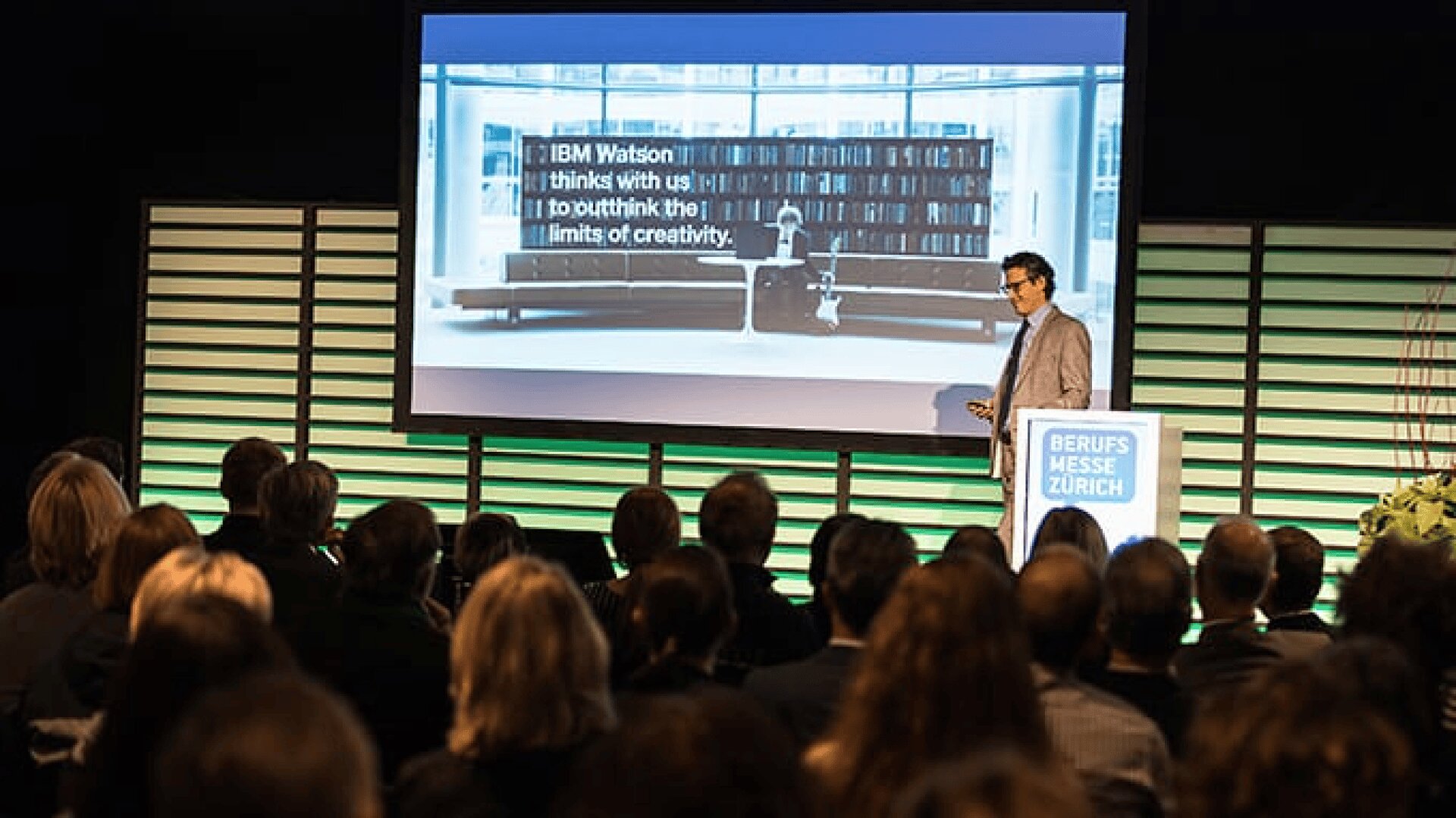 Ein Mann hält einen Vortrag vor einem Publikum. Er steht an einem Podium mit der Aufschrift BERUFS MESSE ZÜRICH, und hinter ihm wird auf einem großen Bildschirm ein IBM Watson-Zitat über Kreativität angezeigt.