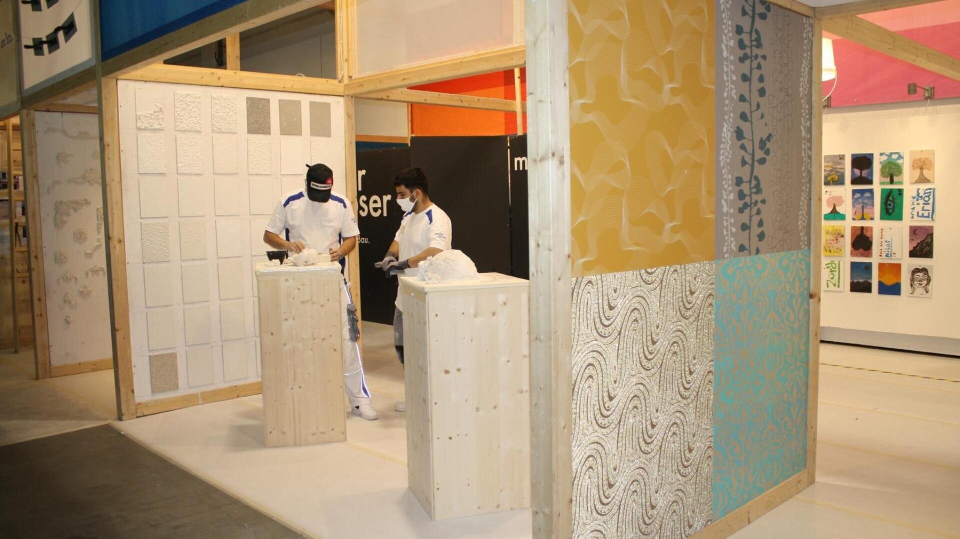 Zwei Personen in weißer Kleidung und mit Gesichtsmasken arbeiten in einem Kunstausstellungsraum mit Holzstrukturen, strukturierten Paneelen und bunten Kunstwerken an den Wänden. Skulpturen sind auf Sockeln ausgestellt.