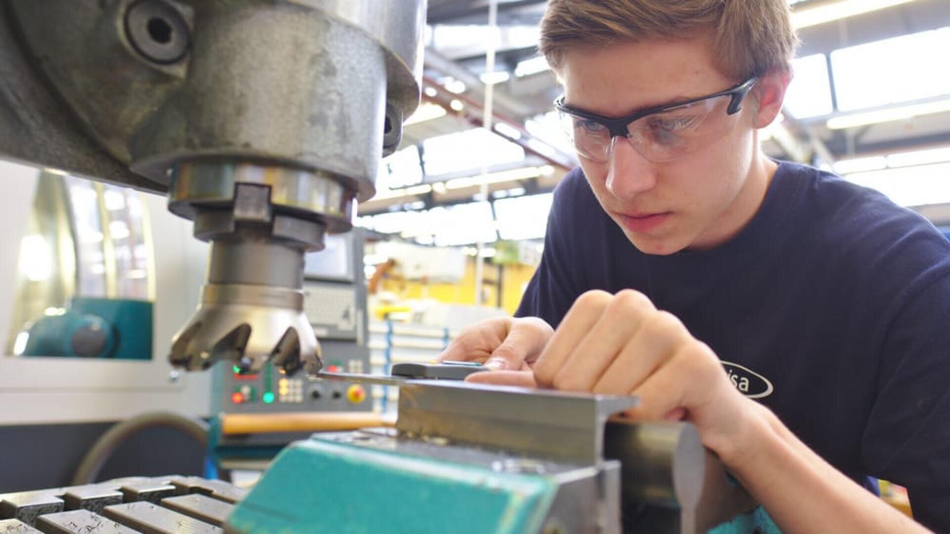 Ein junger Mann mit Schutzbrille arbeitet in einer Werkstatt an einer Fräsmaschine und justiert konzentriert ein in einem Schraubstock eingespanntes Metallteil.