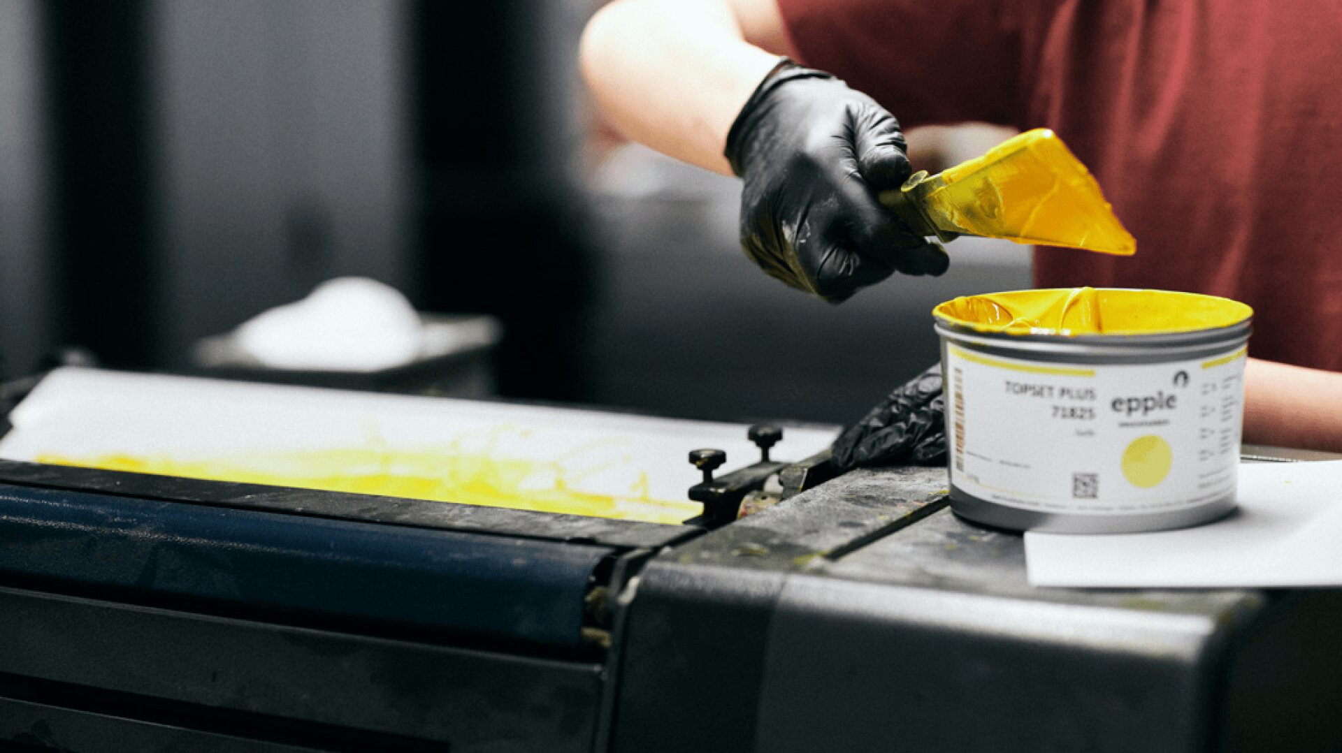 Eine Person mit schwarzen Handschuhen schöpft mit einem Spatel gelbe Tinte aus einer mit "epple" beschrifteten Dose auf eine Druckerpresse.