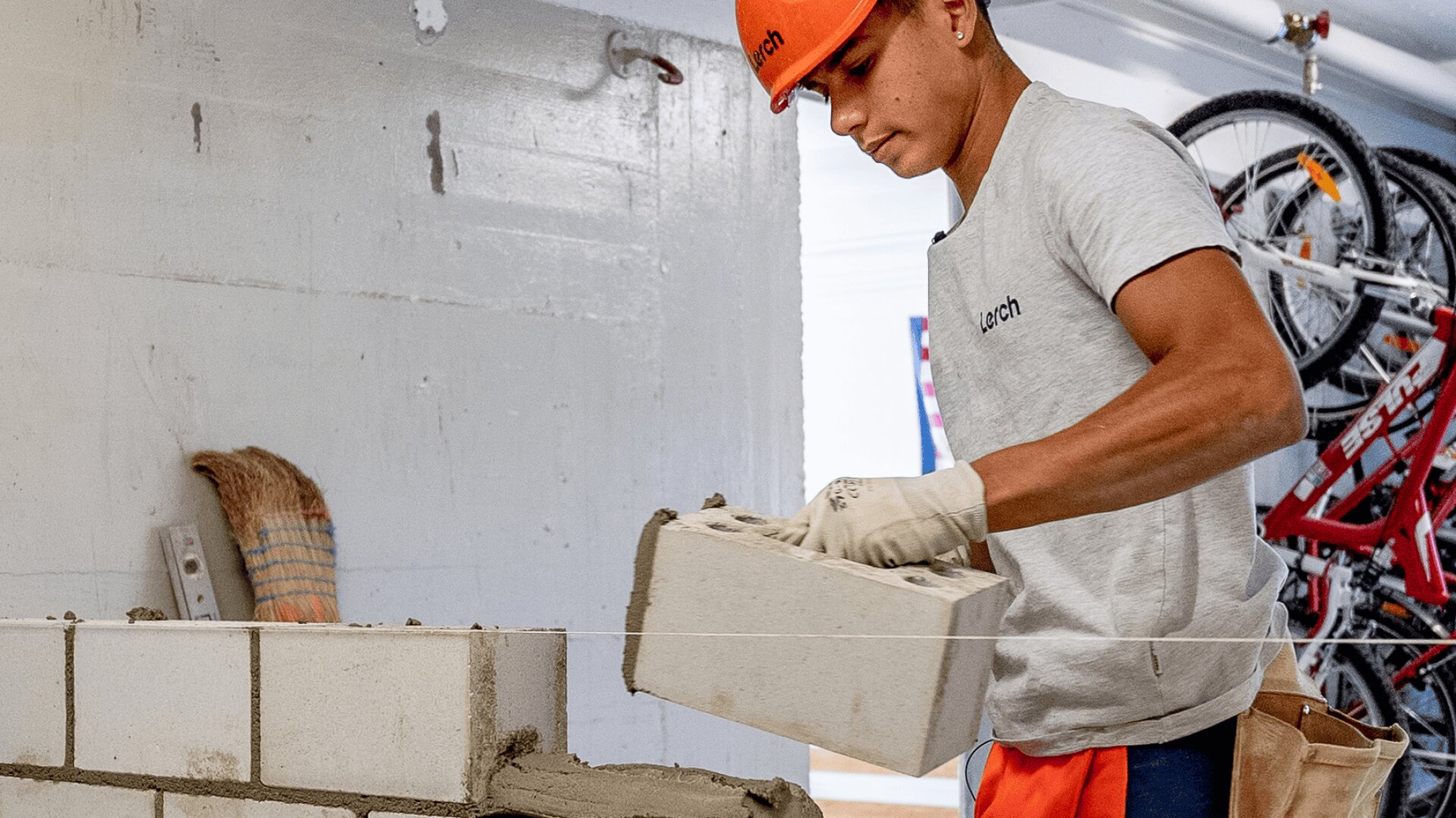 Ein Bauarbeiter im grauen Hemd und mit orangefarbenem Schutzhelm legt einen Betonblock auf eine teilweise errichtete Mauer in einem Gebäude. Er trägt Handschuhe, und im Hintergrund sind Fahrräder zu sehen.