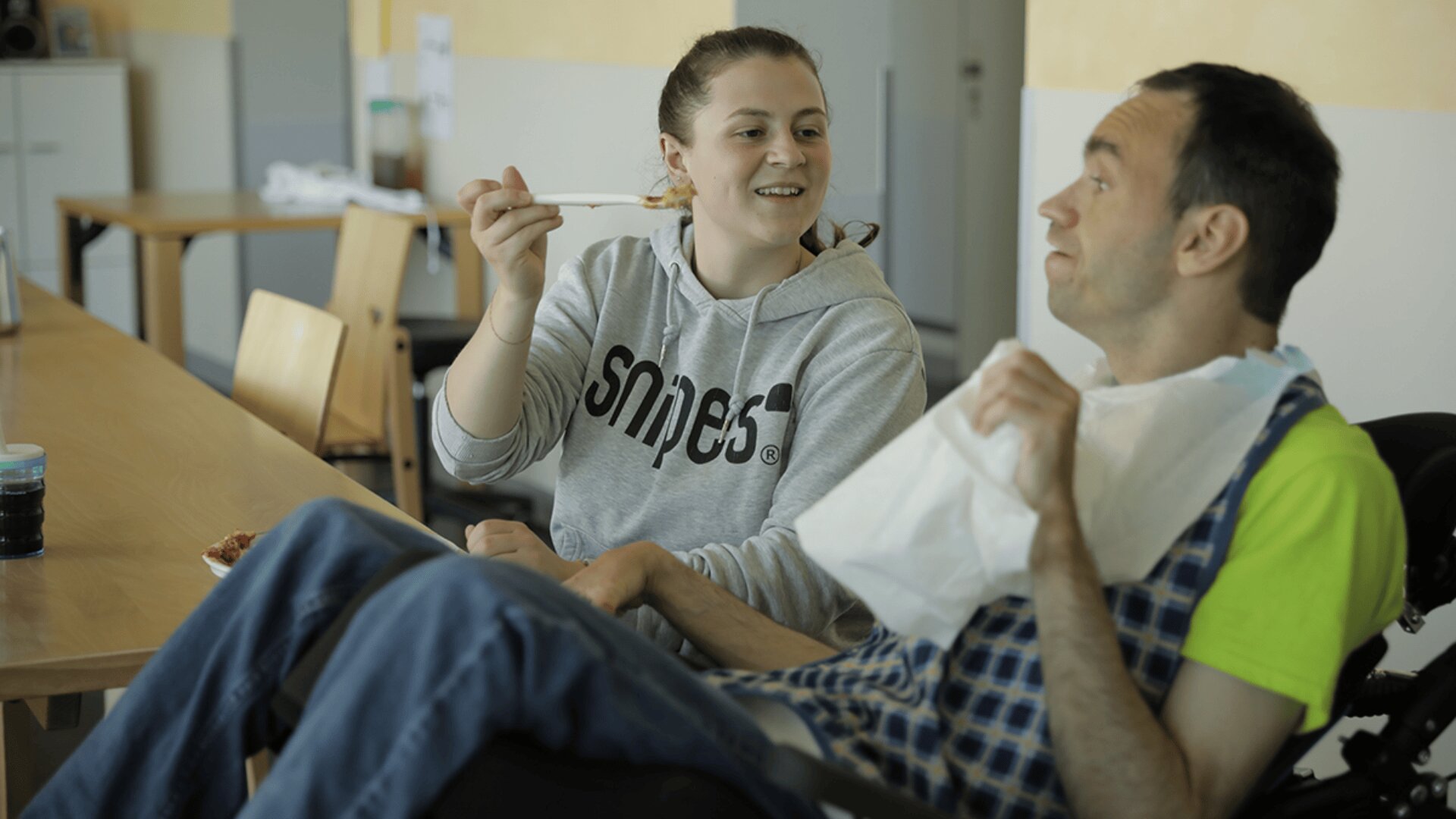 Eine Frau in einem grauen Kapuzenpulli lächelt, während sie einem Mann im Rollstuhl, der ein gelbes Hemd und eine karierte Schürze trägt, beim Füttern hilft und ihm eine Serviette vor den Mund hält. Sie scheinen sich in einem Essbereich zu befinden.