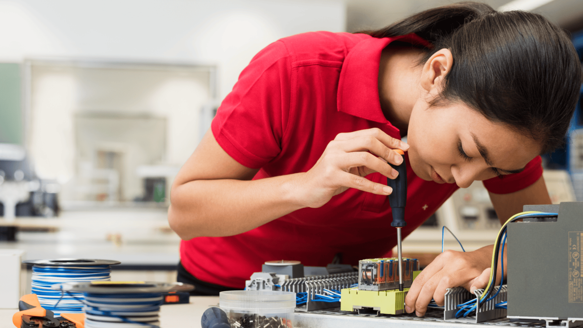 Eine Person in einem roten Hemd arbeitet mit einem Schraubenzieher an Komponenten eines elektronischen Geräts an einem Schreibtisch, umgeben von Kabeln und elektronischen Teilen.