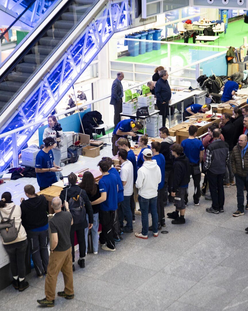 Eine Gruppe von Menschen steht in einer Schlange an einem belebten Anmeldeschalter in der Nähe einer Rolltreppe in einem modernen Gebäude, während Mitarbeiter in blauen Hemden den Teilnehmern helfen.