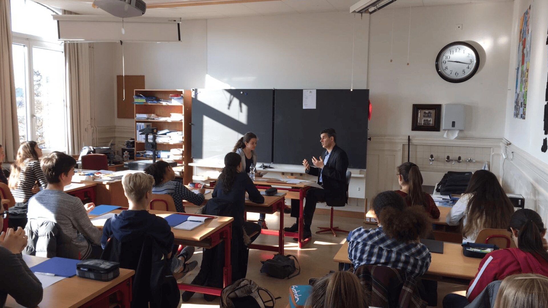 Ein Klassenzimmer mit Schülern, die an den Tischen sitzen und einem Lehrer und einem anderen Schüler gegenübersitzen, die eine Diskussion führen. Das Sonnenlicht fällt durch die Fenster, und eine Uhr an der Wand zeigt an, dass es fast 11:00 Uhr ist.