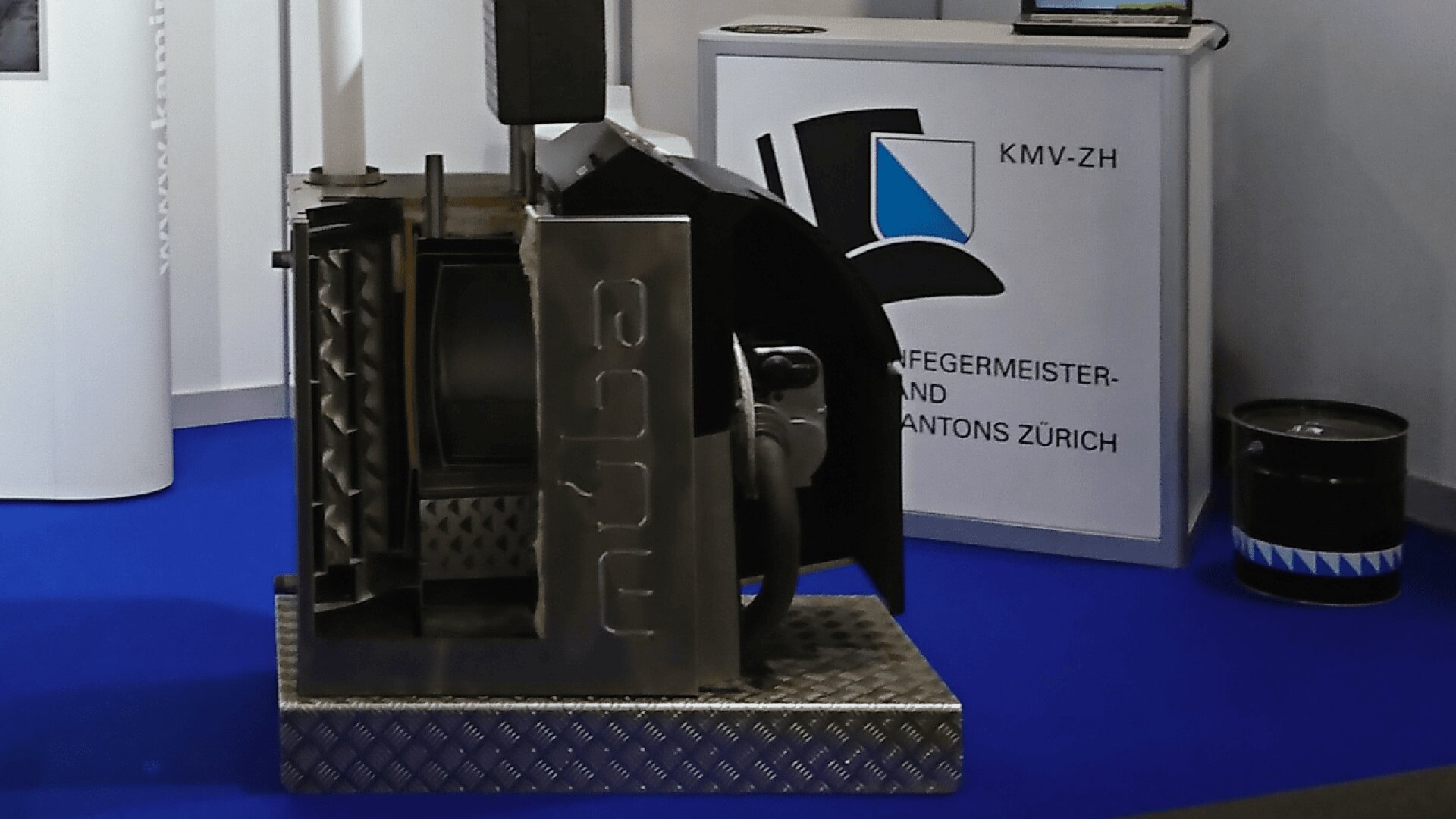 Eine metallene Industriemaschine mit einer Müba an der Seite steht auf einer schachbrettartigen Plattform. Im Hintergrund befindet sich ein Schild mit einem blau-schwarzen Hutlogo und deutschem Text auf einem Tresen. Der Boden besteht aus blauem Teppich.