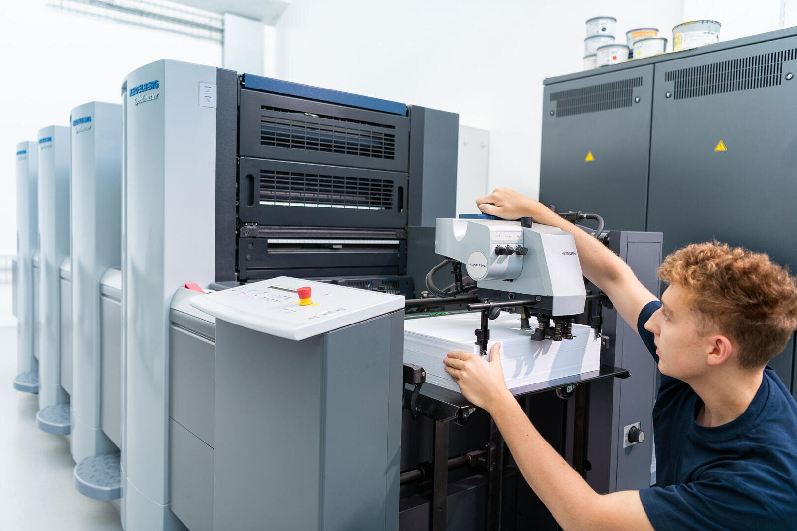 In einer hell erleuchteten, modernen Druckerei bedient eine Person eine große industrielle Druckmaschine und justiert die Papierbögen im Zufuhrfach.