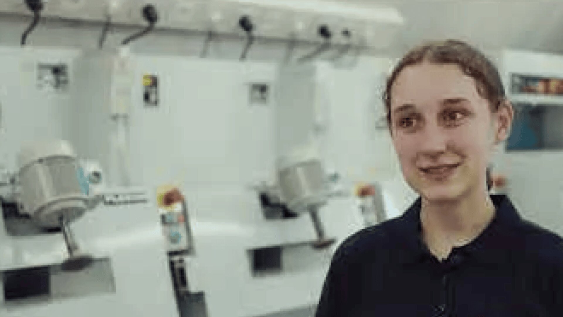 Eine junge Frau mit lockigem Haar und dunklem Hemd steht lächelnd vor weißen Industriemaschinen in einer hell erleuchteten Umgebung.