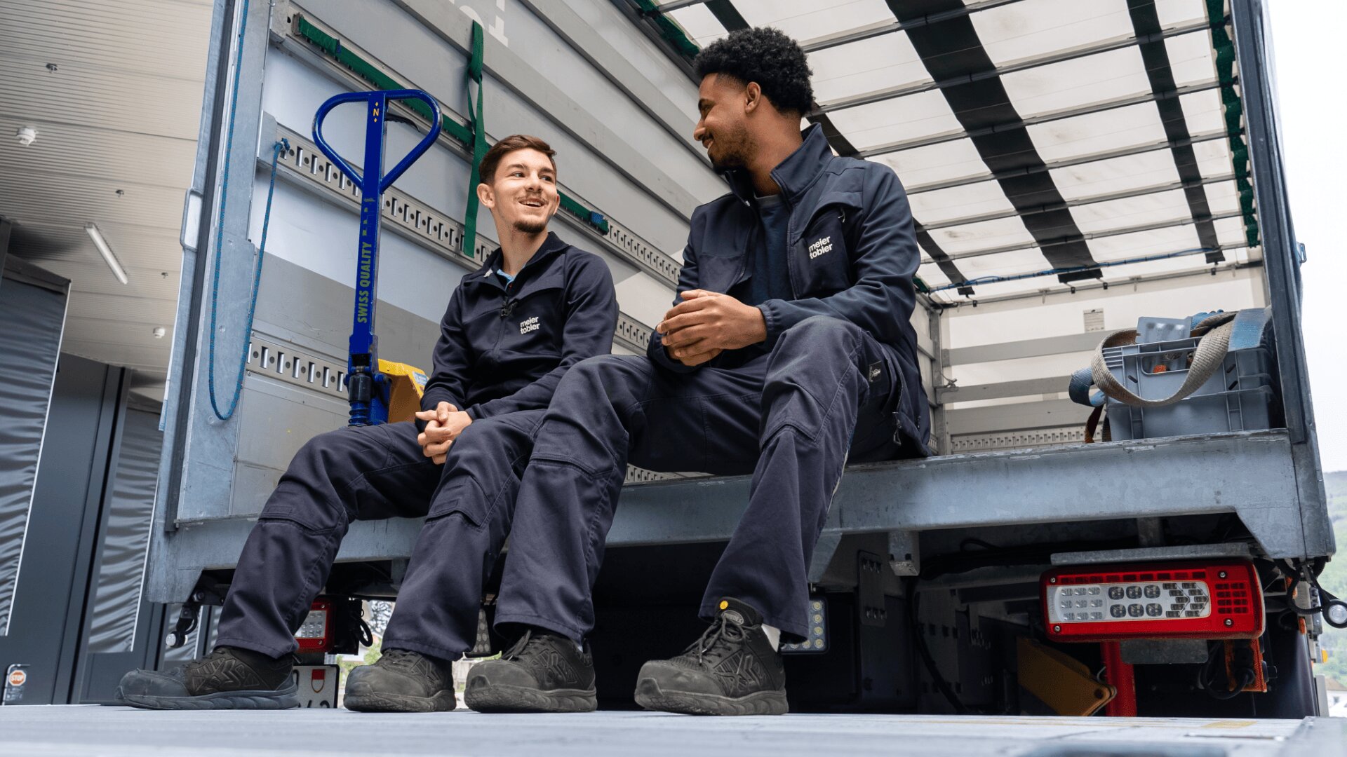 Zwei Männer in Arbeitsuniformen sitzen und unterhalten sich auf der offenen Ladefläche eines Lieferwagens. Sie wirken entspannt und lächeln, während im Lkw Transportgeräte und Werkzeuge zu sehen sind.