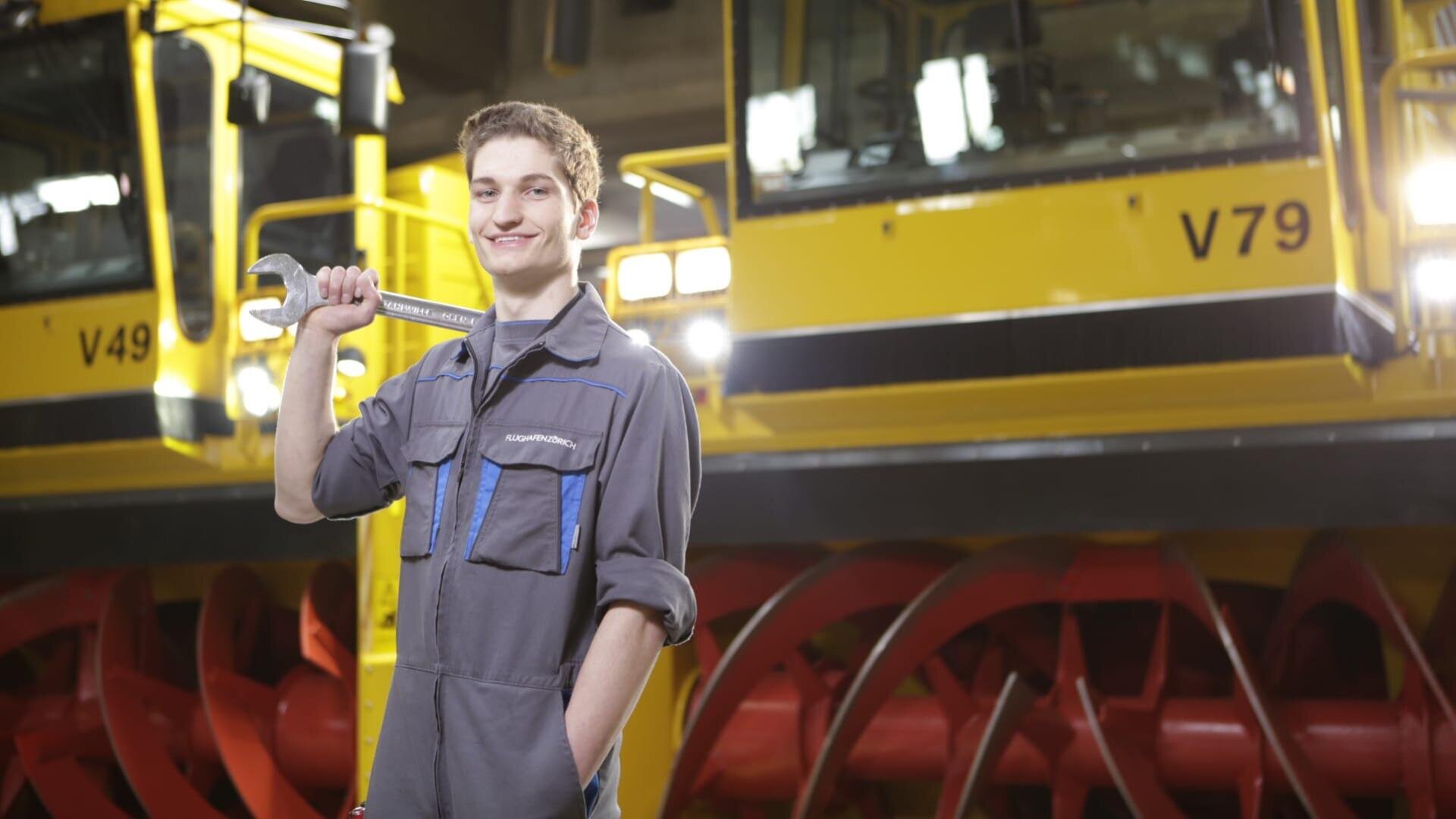 Ein junger Mann in einem grauen Arbeitsoverall hält einen großen Schraubenschlüssel über die Schulter und lächelt. Er steht vor einer leuchtend gelben Industriemaschine mit roten Bauteilen und den sichtbaren Aufschriften V49 und V79.
