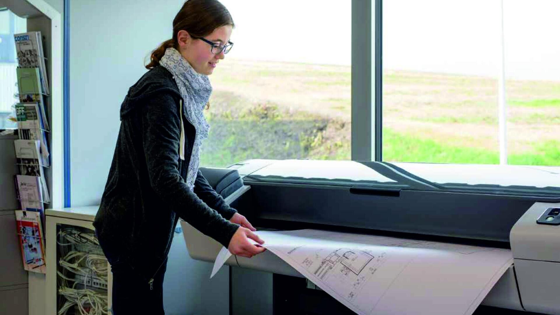 Eine Person mit Brille und Schal steht an einem Großformatdrucker und prüft in einem hellen Raum mit großen Fenstern, die den Blick nach draußen freigeben, einen Bauplan oder eine Architekturzeichnung.