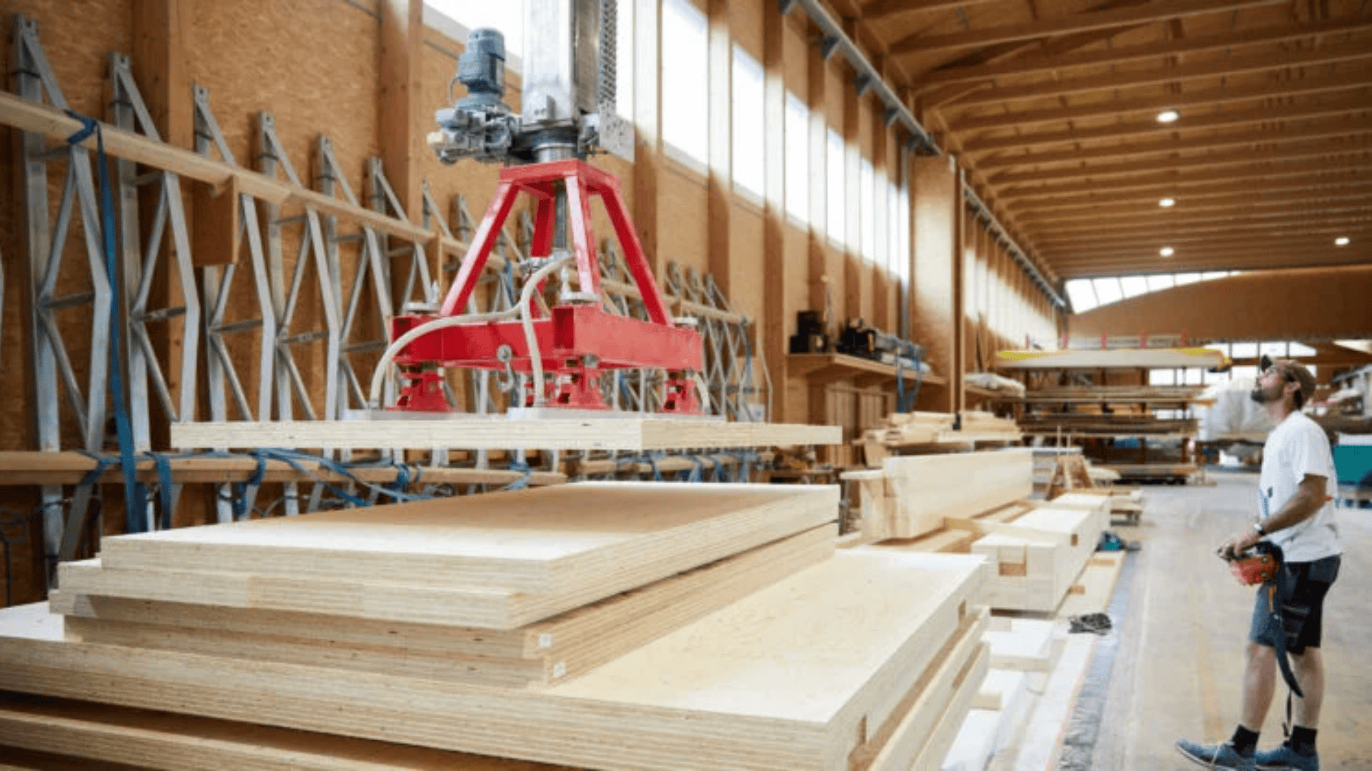 Ein Mann in einer hellen, geräumigen Werkstatt beobachtet, wie ein großer roter Roboterarm Sperrholzplatten auf einen Stapel hebt. Die Werkstatt hat Holzbalken, hohe Decken und Reihen von gelagerten Materialien.