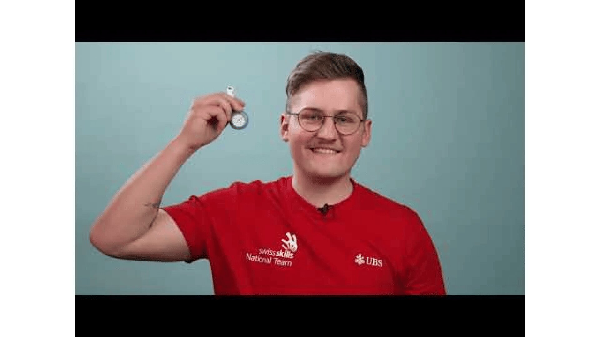 Eine lächelnde Person mit Brille und rotem SwissSkills Nationalmannschaftstrikot hält eine kleine Silbermedaille oder Münze vor einem einfarbig blauen Hintergrund hoch.