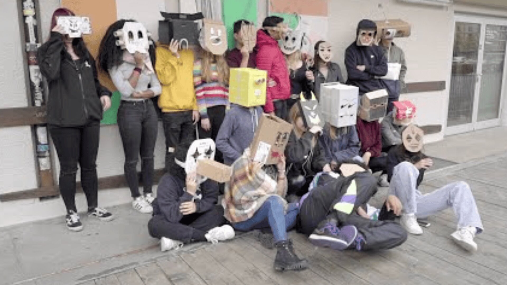 Eine Gruppe von Menschen posiert im Freien und trägt verschiedene selbstgemachte Masken und Kopfbedeckungen aus Pappkartons und Papier, einige mit gezeichneten oder ausgeschnittenen Gesichtern. Sie sind leger gekleidet und scheinen sich zu amüsieren.