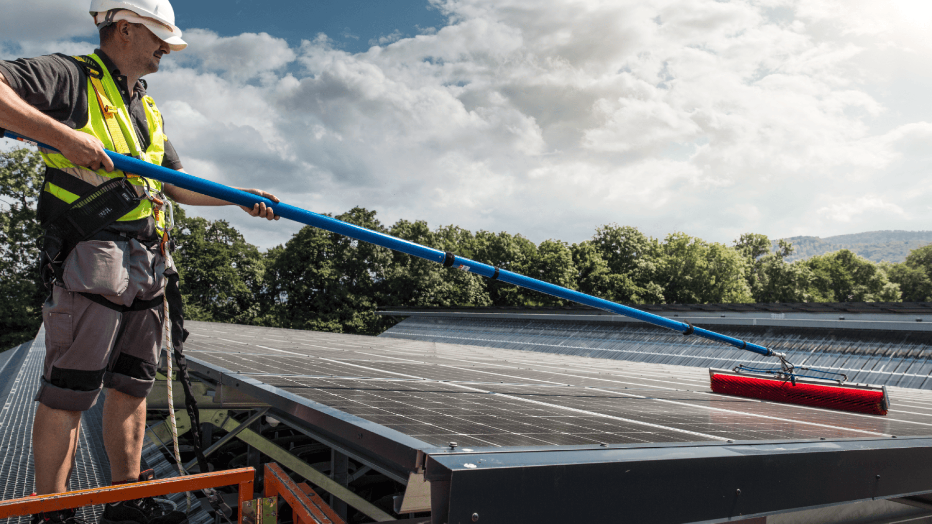 Ein Arbeiter in Schutzkleidung steht auf einer Plattform und reinigt mit einer langen Bürste Solarpaneele auf einem Dach unter einem teilweise bewölkten Himmel mit Bäumen im Hintergrund.