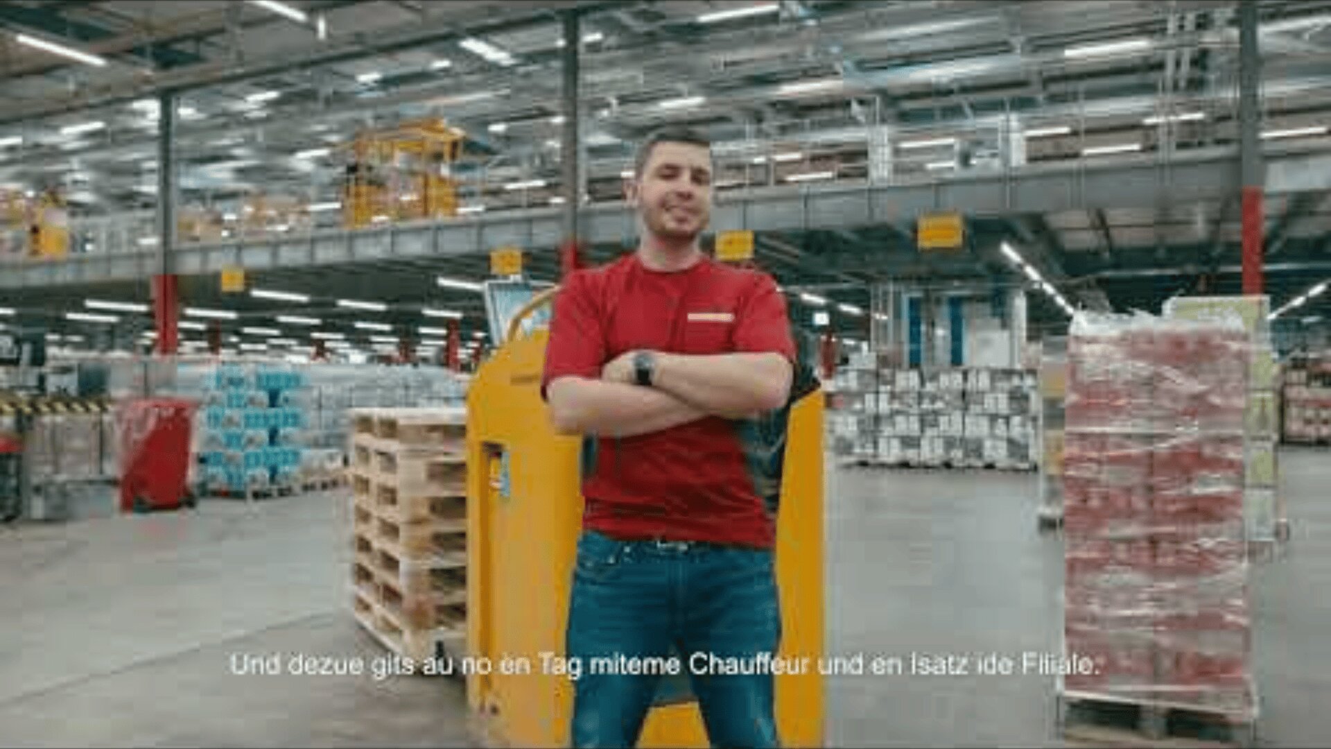 Ein Mann in einem roten Hemd steht mit verschränkten Armen in einer großen Lagerhalle, umgeben von Paletten und Kisten. Oberlichter erhellen die industrielle Umgebung. Am unteren Rand des Bildes erscheinen deutsche Untertitel.