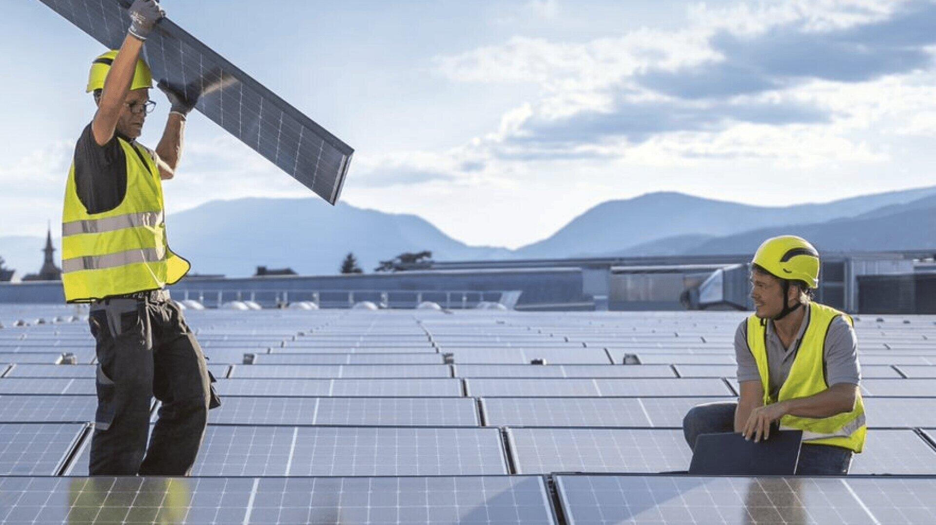 Zwei Arbeiter in gelben Sicherheitswesten und mit Helmen installieren Solarzellen auf einem Dach, mit Bergen und einem teilweise bewölkten Himmel im Hintergrund.