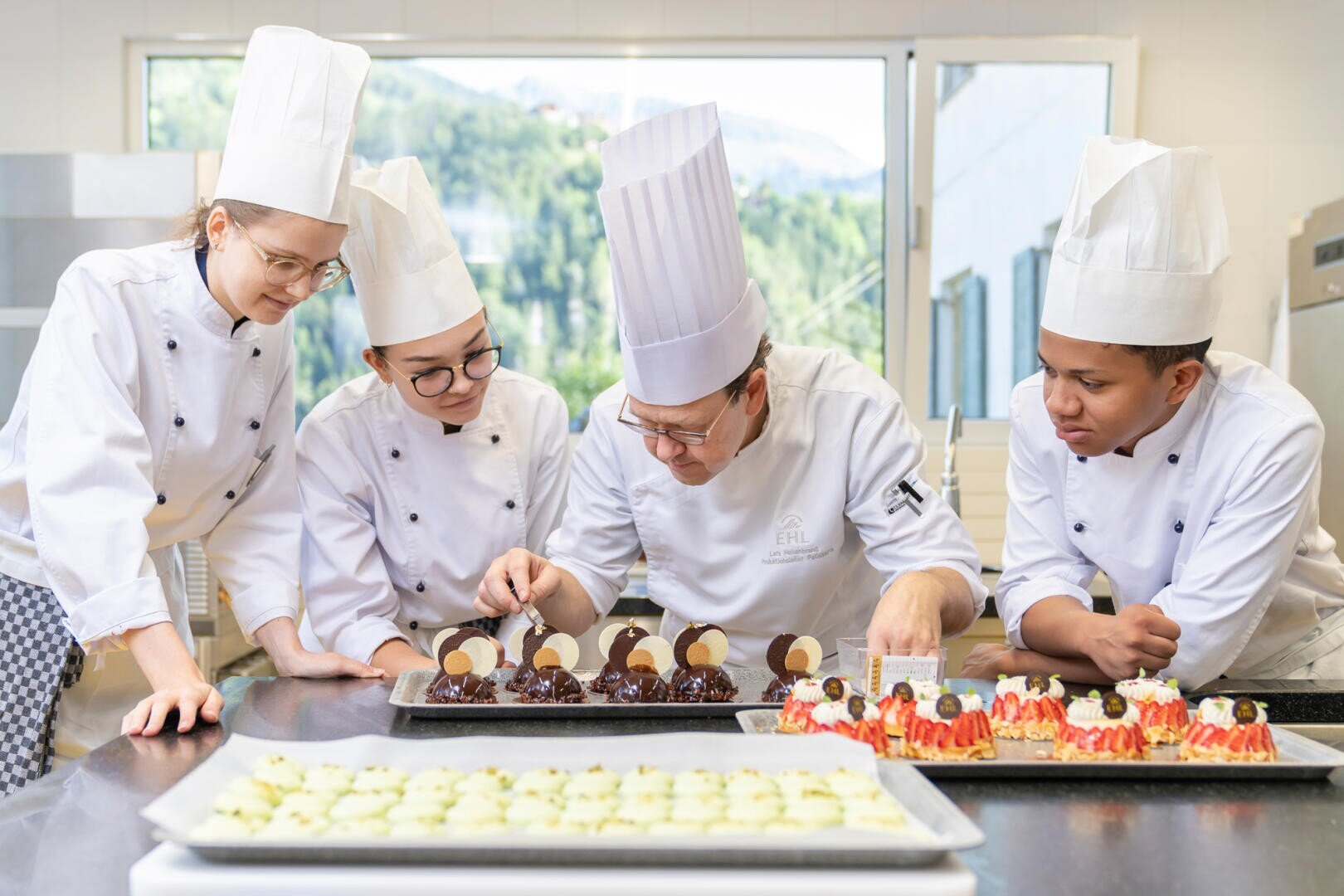 Vier Konditoren in weißen Uniformen und Mützen sind um eine Küchentheke versammelt und beobachten aufmerksam, wie einer der Köche Schokoladendesserts auf einem Tablett dekoriert. Andere fertige Desserts sind auf dem Tresen zu sehen. Große Fenster geben den Blick auf eine malerische Landschaft frei.