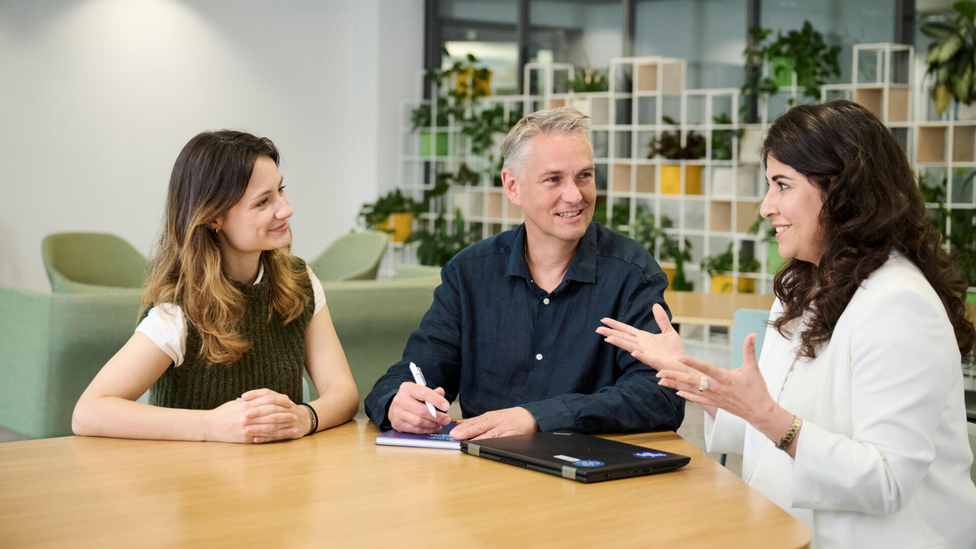 Drei Personen sitzen an einem runden Tisch in einem modernen Büro und führen eine Diskussion. Eine Person gestikuliert beim Sprechen, eine andere lächelt, und eine dritte macht sich Notizen. Im Hintergrund stehen Pflanzen und Regale.