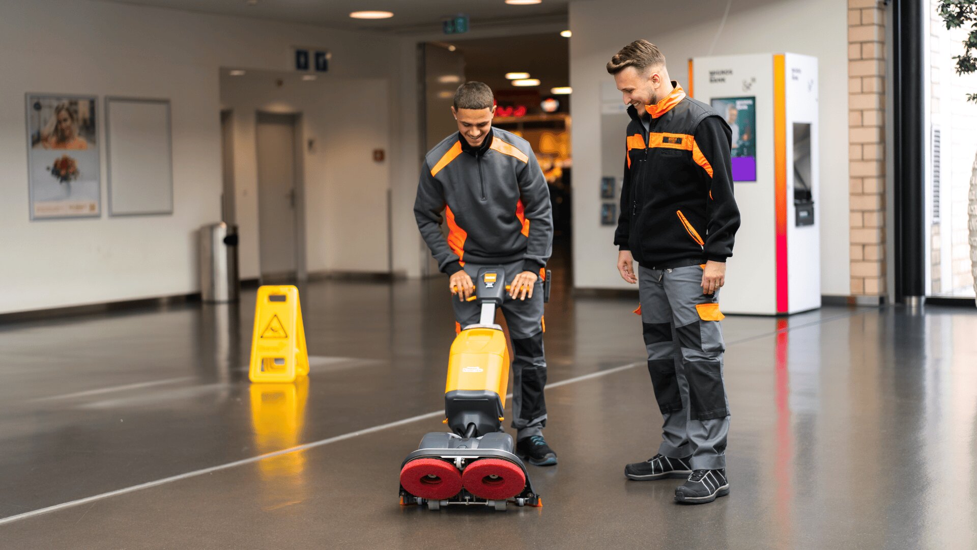 Zwei Männer in Arbeitsuniformen reinigen in einem modernen Gebäude einen glänzenden Boden mit einer handelsüblichen Scheuersaugmaschine. Einer bedient die Maschine, während der andere zuschaut. Im Hintergrund ist ein gelbes Schild mit der Aufschrift "Vorsicht nasser Boden" zu sehen.