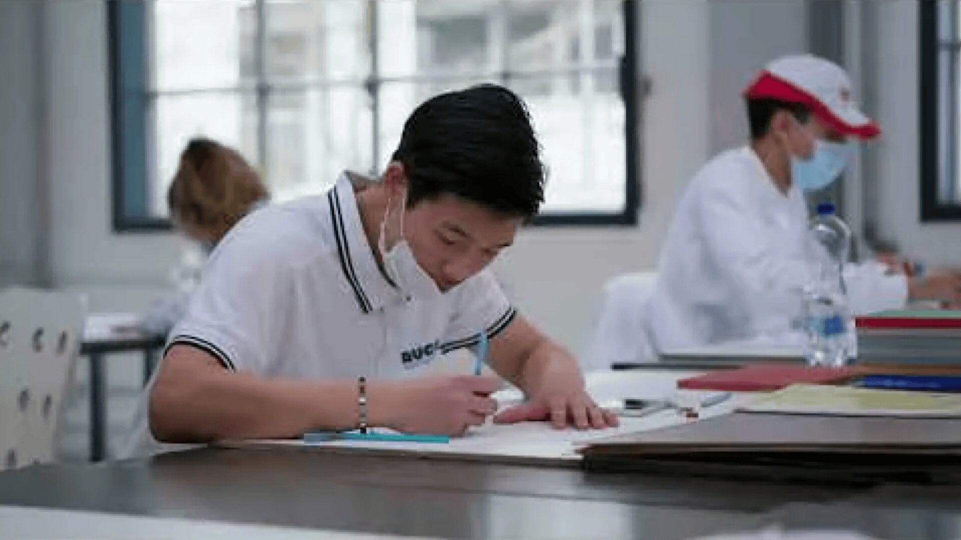 Ein Schüler, der eine Maske trägt, schreibt an einem Schreibtisch in einem Klassenzimmer in ein Notizbuch, während andere maskierte Schüler im Hintergrund sitzen und arbeiten.
