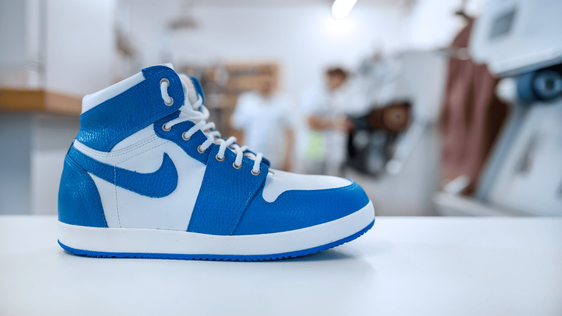 Eine Nahaufnahme eines blauen und weißen Nike High-Top-Sneakers, der auf einer weißen Fläche ausgestellt ist, mit einem unscharfen Hintergrund, der Menschen und Geräte in etwas zeigt, das eine Werkstatt oder ein Geschäft zu sein scheint.