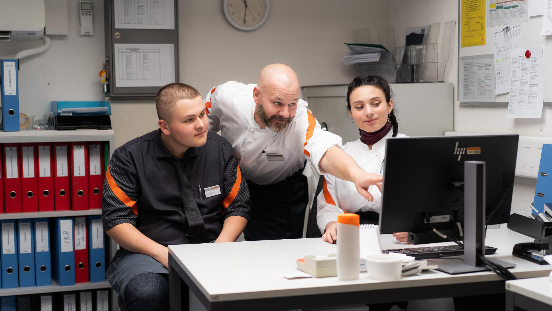 Drei Kollegen in Uniform sitzen um einen Schreibtisch in einem Büro und schauen auf einen Computerbildschirm. Eine Person deutet auf den Bildschirm, während die anderen aufmerksam zusehen. Im Hintergrund sind Regale und Dokumente zu sehen.