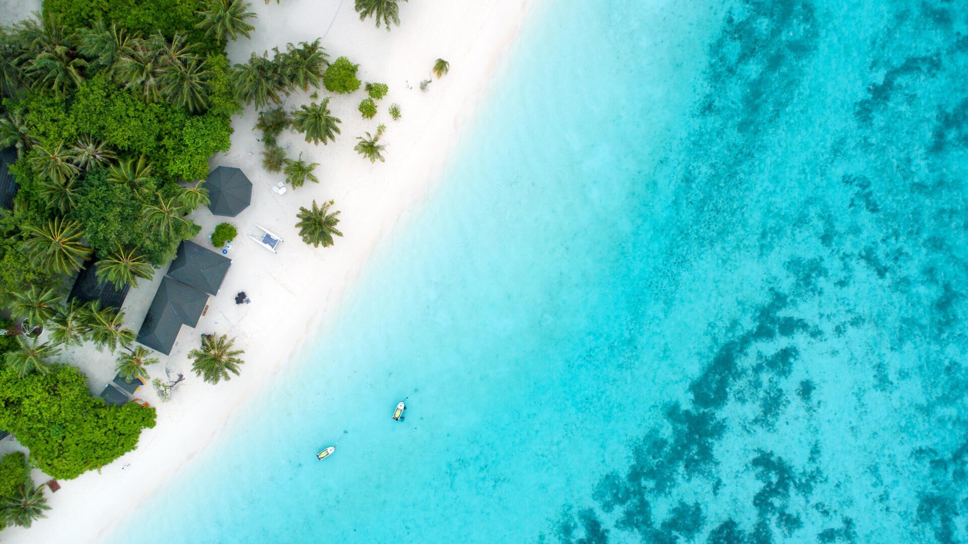 Luftaufnahme eines tropischen Strandes mit üppigen grünen Bäumen, weißem Sand und türkisfarbenem Wasser. Zwei kleine Boote schwimmen in der Nähe des Ufers, und einige Gebäude sind zwischen den Palmen eingebettet.