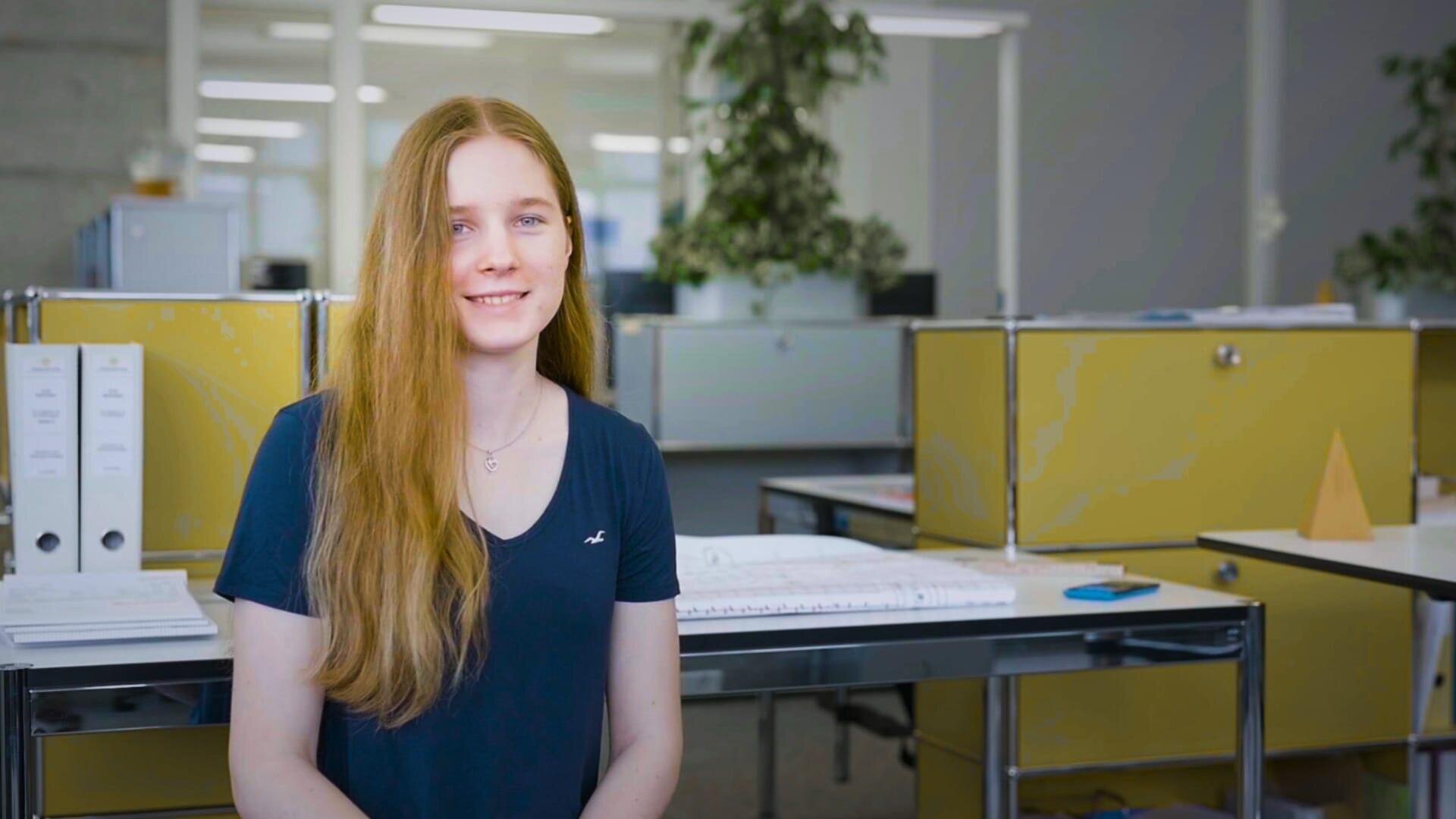 Eine junge Frau mit langen roten Haaren und einem marineblauen Hemd sitzt lächelnd in einem modernen Büro mit gelben Schränken, Pflanzen und Büromaterial im Hintergrund.