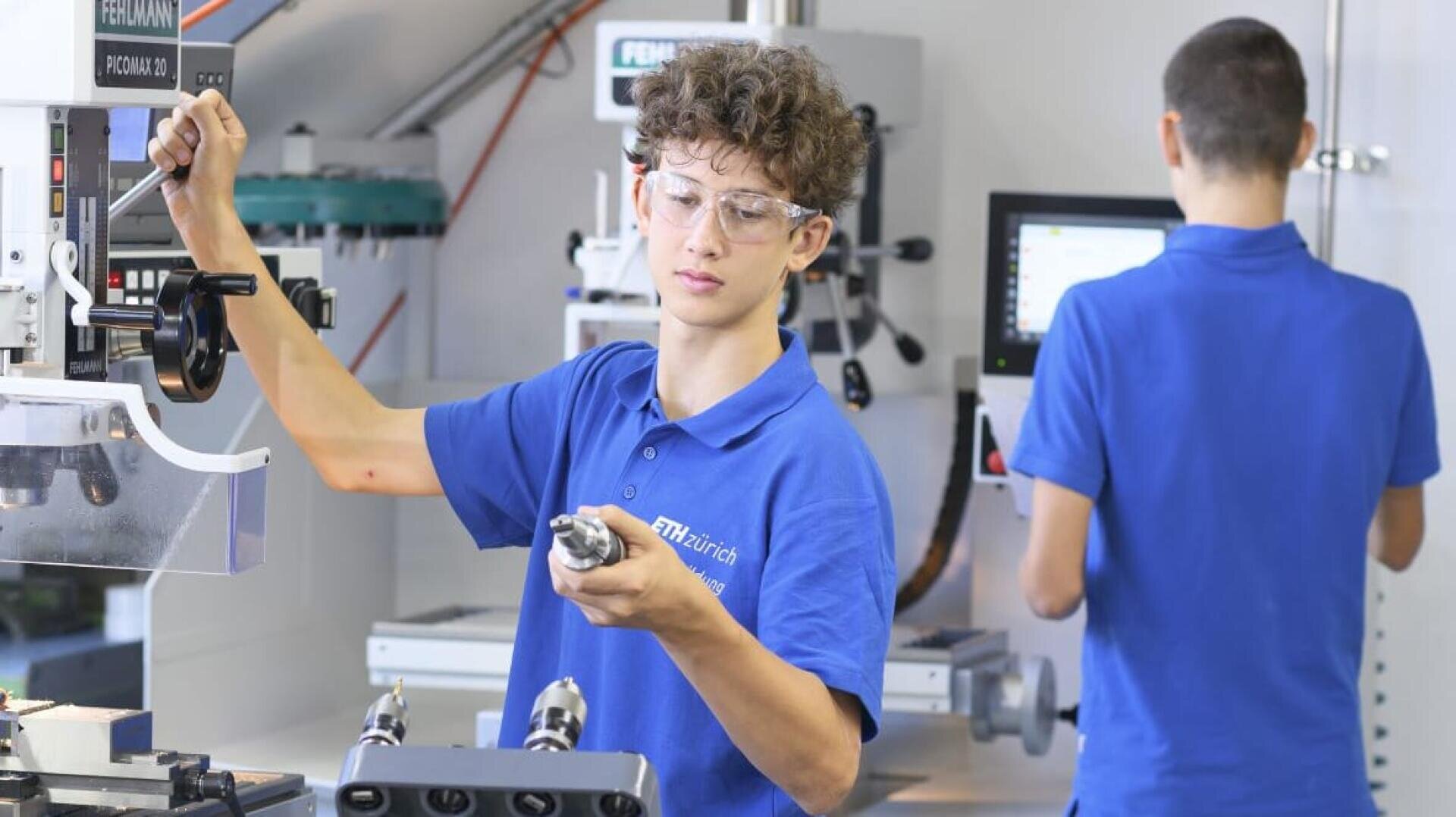 Ein Jugendlicher mit Schutzbrille und blauem Polohemd bedient eine Maschine in einer Werkstatt und hält ein Metallteil. Ein anderer Schüler in einem blauen Hemd arbeitet an einem Computer oder einer Maschine im Hintergrund.