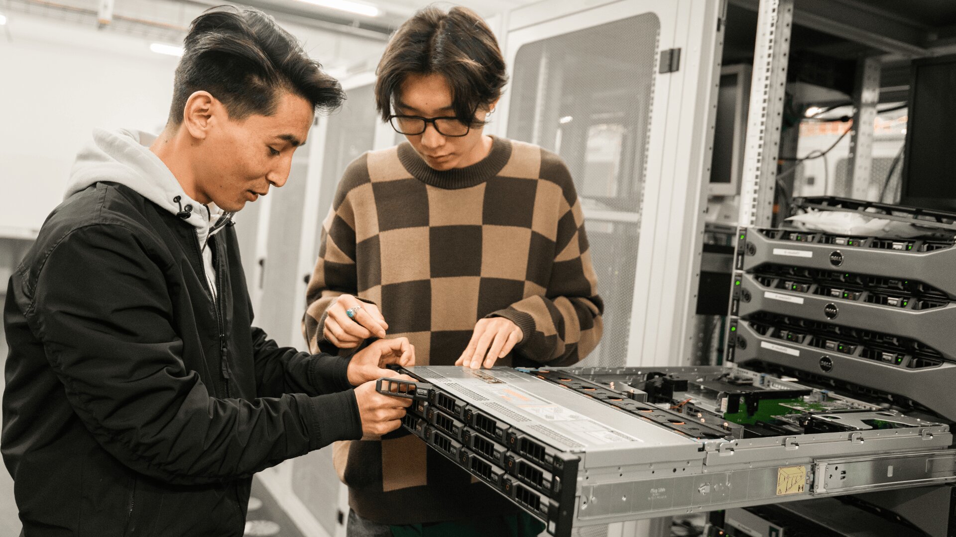 Zwei Personen arbeiten zusammen, um einen Server in einem Rechenzentrum zu installieren oder zu reparieren, wobei die Serverschränke und die Computerausrüstung um sie herum sichtbar sind. Einer hält ein Werkzeug, während der andere einen Teil des Servers ausrichtet.