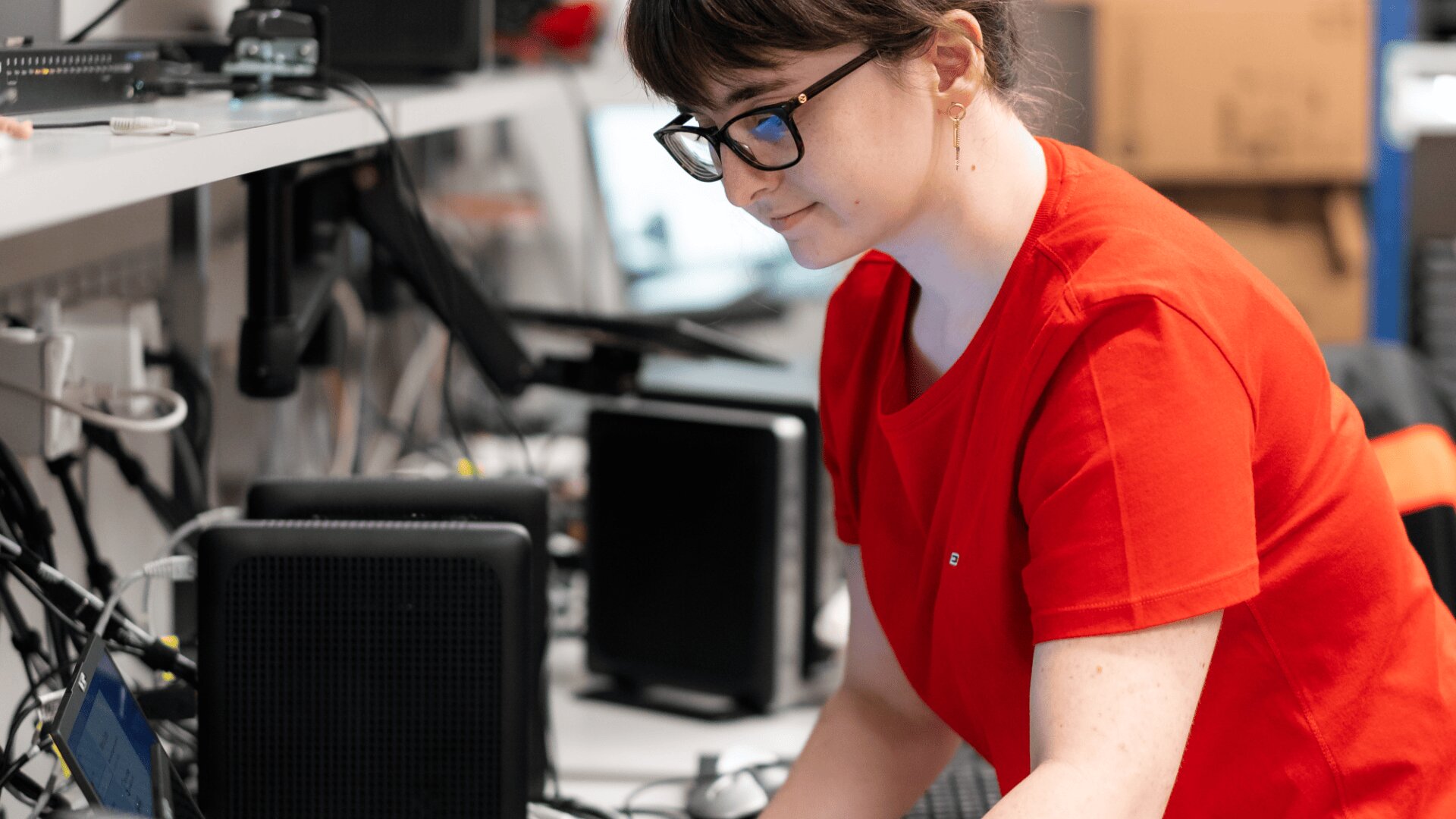 Eine Person mit Brille und rotem Hemd arbeitet an einem Schreibtisch mit verschiedenen elektronischen Geräten und Kabeln und konzentriert sich auf eine Aufgabe in einem technischen Arbeitsbereich.